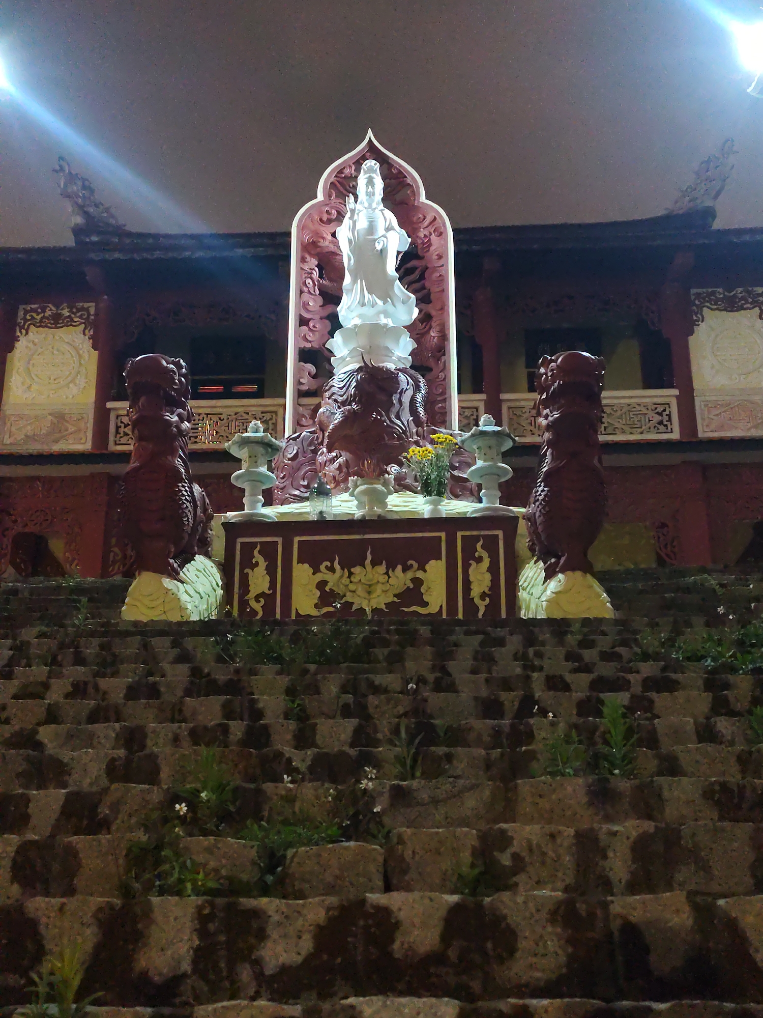 The image depicts a nighttime scene at a temple or shrine in Da Lat, Vietnam. A large set of stone steps leads up to a platform where a white statue of a deity, possibly Quan Am, stands. The statue is set within a carved, ornate alcove, and two decorative dragon statues flank the platform. On the platform there's what appears to be an offering table, with flowers and lanterns. The architecture of the building behind the statue exhibits traditional Vietnamese design elements. The lighting is somewhat dim, with an artificial light source illuminating the statue and the building. Plants are growing between the stone steps.