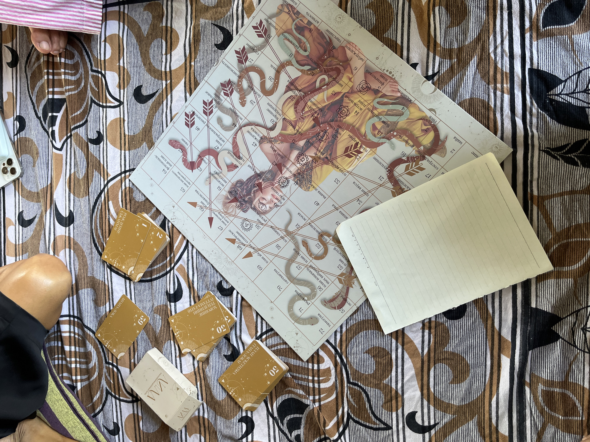 The image shows a close-up, top-down view of a board game and several decks of cards laid out on a patterned fabric. The game board features a central image of a woman entwined with snakes, surrounded by numbered spaces with Russian text. Several stacks of square, gold-colored cards with similar designs are placed to the left of the board, along with one rectangular white box that also contains cards. A blank, lined notepad is positioned in the lower right quadrant of the image, partially covering the game board. The fabric beneath the objects has a repeating pattern of brown and black wavy lines interspersed with thin white lines. In the upper portion of the image, a person's bare leg and knee, along with a black garment, are visible, suggesting they are seated or lying on the fabric. The lighting is consistent with natural daylight. The specific location in Weligama, Sri Lanka, cannot be determined from the visual information present.