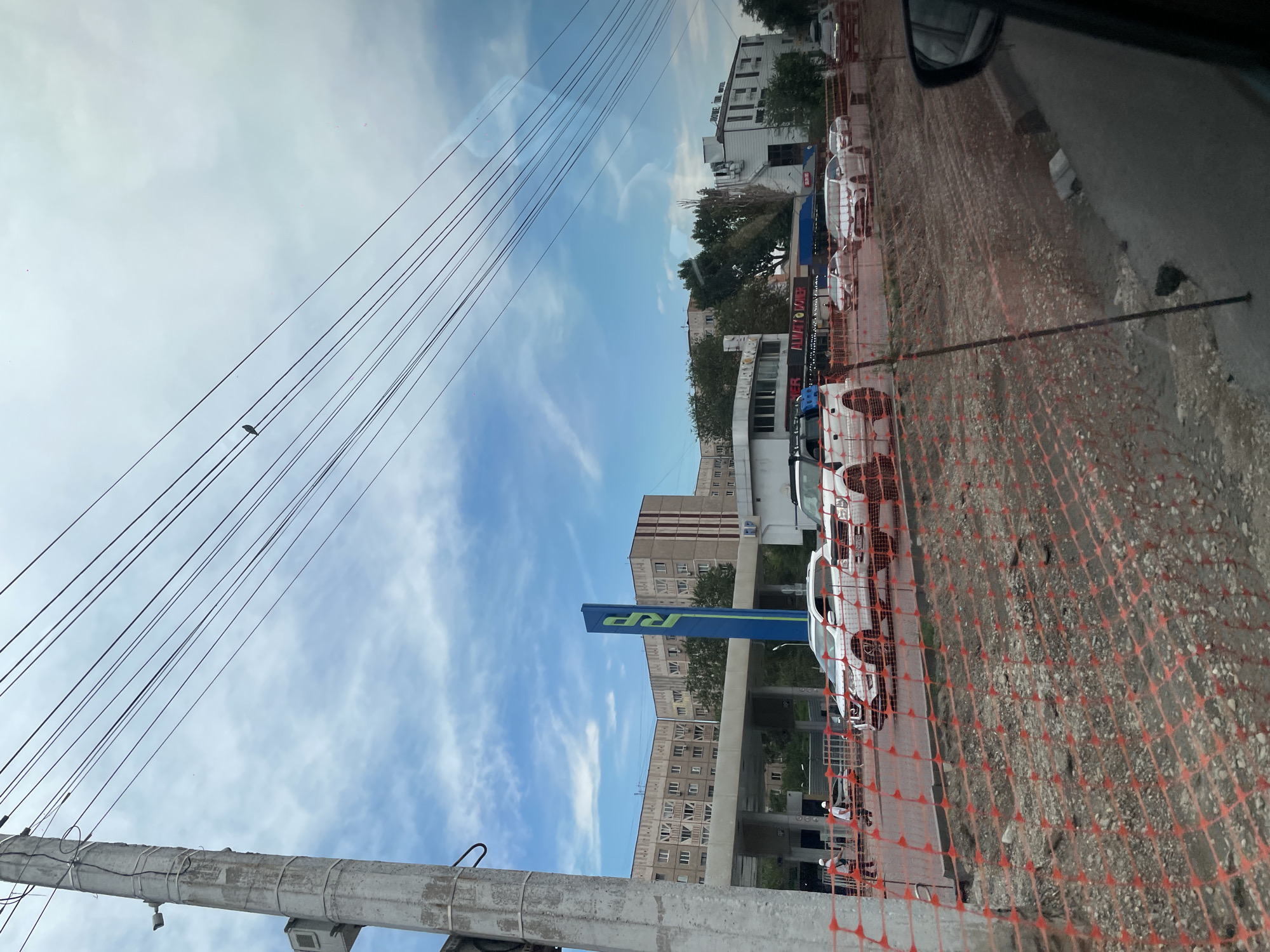 The image depicts a street scene in Almaty, Kazakhstan, likely during daytime under a cloudy sky. In the foreground, a section of road is under construction, marked by orange netting and piles of gravel. Several white vehicles, including SUVs and sedans, are visible on the road. To the left, a concrete utility pole with electrical wires stretches across the frame.
In the background, a gas station with a prominent blue and green "RP" sign is situated. Adjacent to it are multi-story apartment buildings and other commercial establishments, including a building with "HOME INN HOTEL" signage. Two individuals wearing white hard hats and vests are visible near the gas station, possibly construction workers. The overall impression is that of an active urban environment with ongoing infrastructure work.