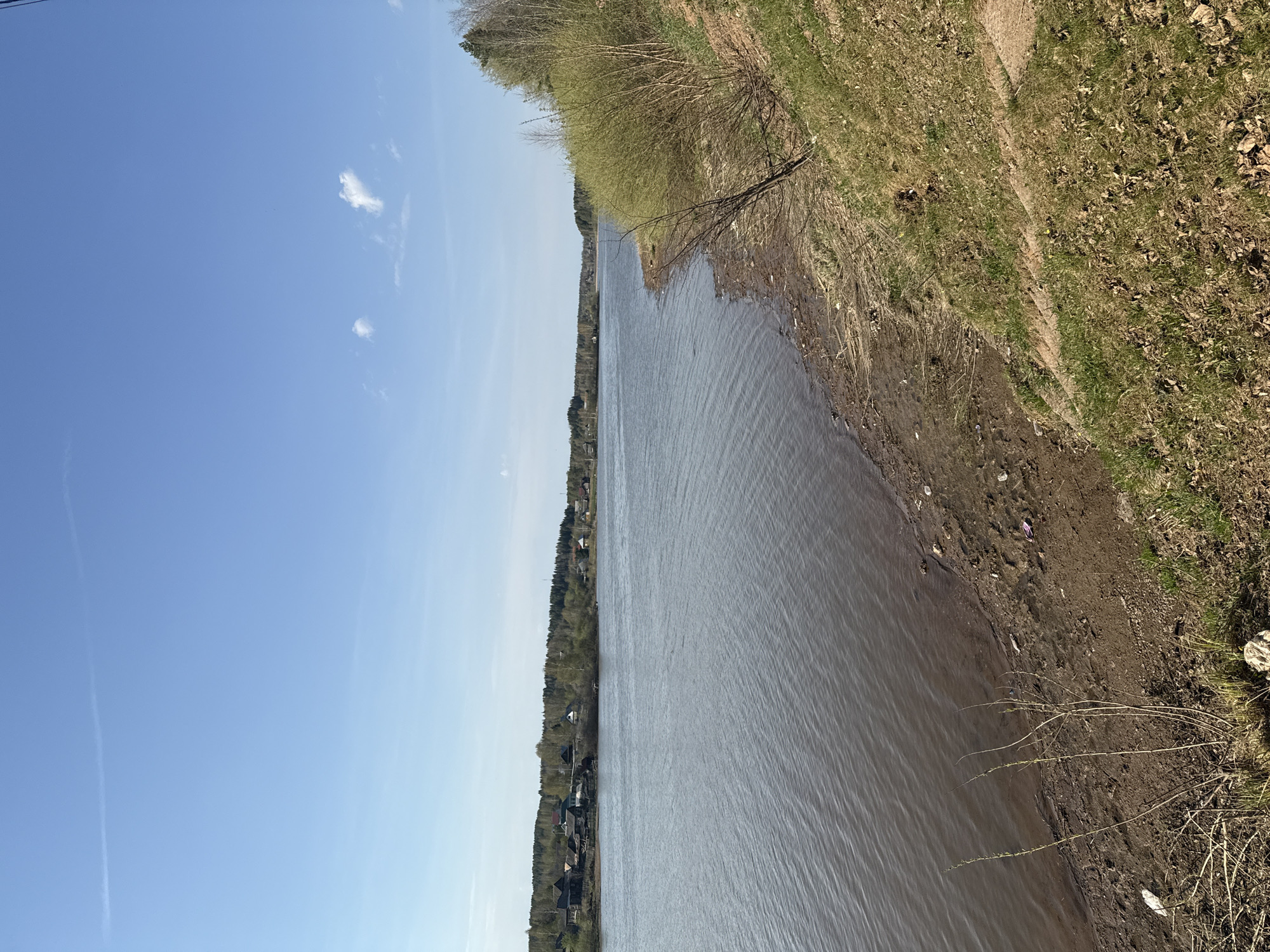 The image shows a body of water, likely a river or lake, with a grassy bank on the right side. The water is reflecting the sky, which is a clear blue with a few small clouds. On the far side of the water, there are trees and buildings. The location is most likely Luzhkovo, Russia.