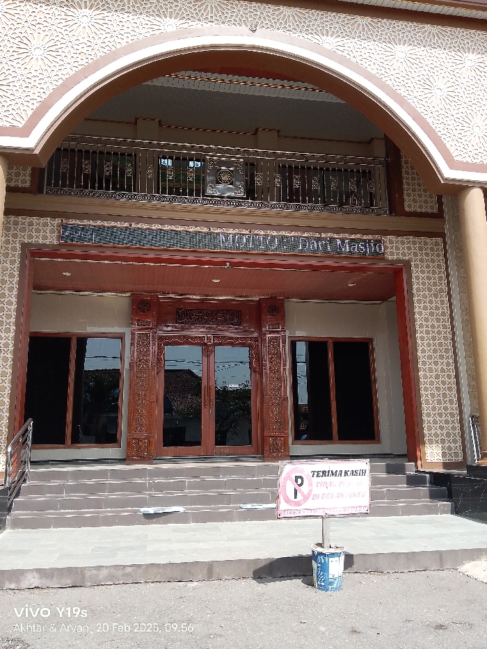 This daylight image captures the exterior of a mosque or Islamic center in Patrol, Indonesia, on February 20, 2025, at 09:56 AM. The building exhibits traditional Islamic architecture with a large, decorative archway forming the main entrance. The facade is adorned with intricate white and beige geometric patterns.
Through the arch, a second-story balcony with a detailed metal railing is visible. Below the balcony, a partial sign reads "MOTTO "Dari Maspo"" or "Masjid". The main ground-level entrance features ornate, carved wooden double doors, flanked by large windows on either side that reflect parts of the surrounding environment.
A wide set of grey steps leads up to the entrance. In front of the steps, a white sign is placed on a post made from a painted bucket. The sign clearly displays a red no-parking symbol. Below it, text in Indonesian reads: "TERIMA KASIH" (Thank You) and "TIDAK PARKIR DI DEPAN PINTU" (No Parking in front of the door). The lighting suggests a clear and sunny day. No people are visible in the scene.