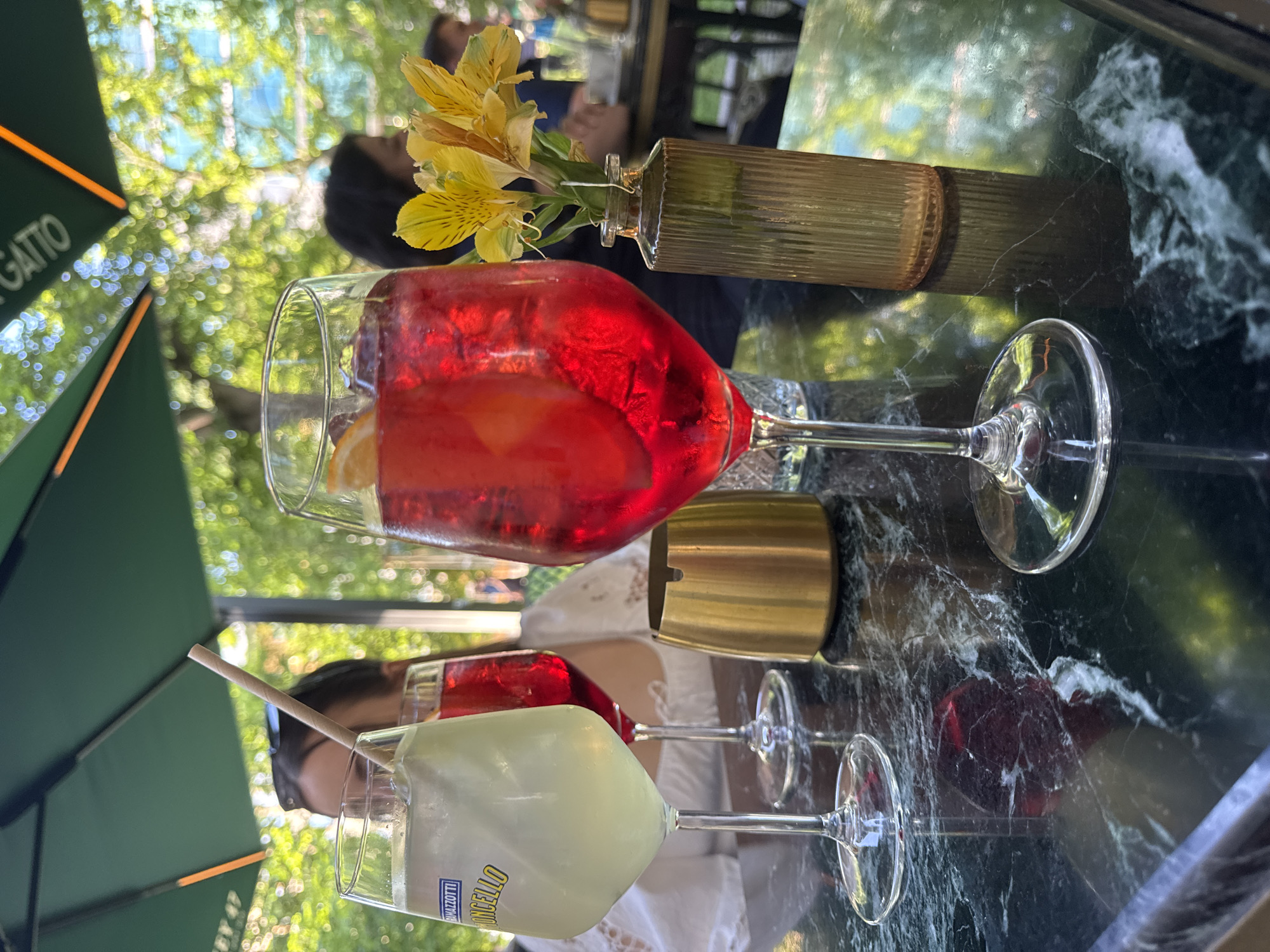 The image depicts a table setting at an outdoor cafe, likely in Sofia, Bulgaria, based on the cafe name "Il Gatto" visible on an umbrella. A woman with dark hair is partially visible in the background. There are two drinks in stemmed glasses; one is a red alcoholic beverage with an orange slice, likely an Aperol spritz. The other is a pale yellow drink, possibly a Limoncello spritz, with a straw. There's a small gold-colored vase containing a few yellow flowers on the table. The table itself has a dark, marbled surface. The scene suggests a casual social gathering at a cafe.
