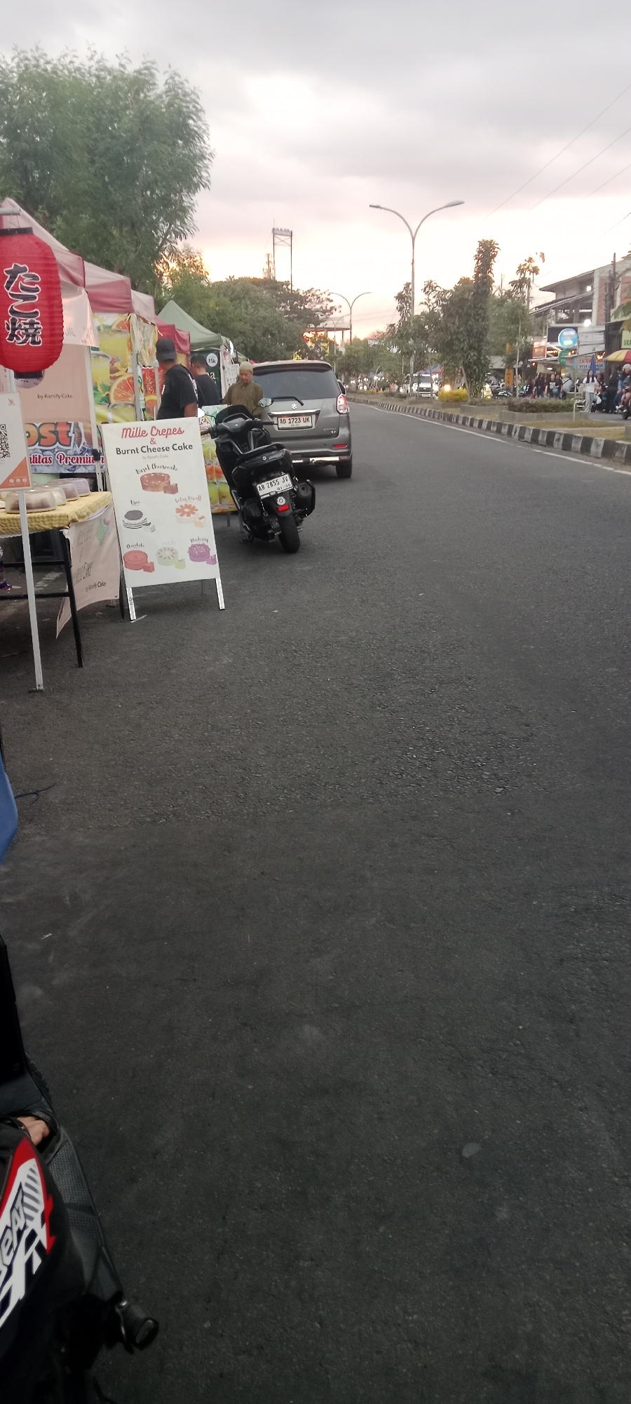 A street scene in Ngemplak, Indonesia features a food stall with a red lantern and a sign for "Mille Crepes & Burnt Cheese Cake". A gray car with license plate AD 1723 UK and a black motorcycle with license plate AD 2855 JV are parked on the side of the road. Several people are visible at the food stall and on the street, some interacting with the stall. In the background, buildings and trees line the street, with a traffic light visible in the distance. The sky is overcast, suggesting late afternoon or early evening.