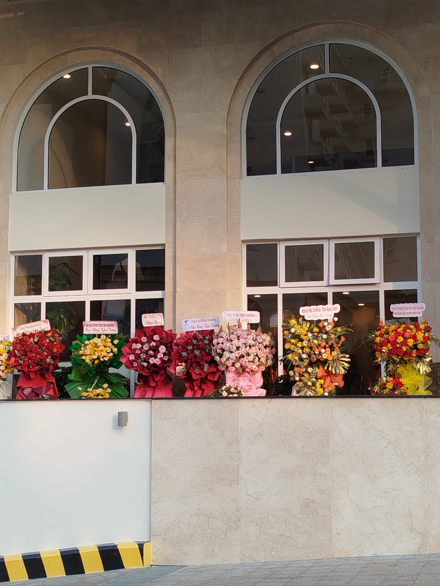 The image shows the facade of a building in Da Lat, Vietnam, likely during a grand opening or celebratory event. The building has two arched windows above rectangular ones, and a light-colored, possibly stone or tiled, exterior. Below the windows, a series of floral arrangements are displayed, each with a sign bearing text in Vietnamese. The floral arrangements are varied in color, with reds, yellows, pinks, and oranges dominating. Each arrangement is wrapped in colorful fabric. The signs contain business names and celebratory messages. A section of wall is visible at the bottom of the frame with a striped warning marking. The building appears to be a commercial establishment or office building. The reflections in the windows suggest a developed area with other buildings nearby. The light suggests late afternoon or early morning.