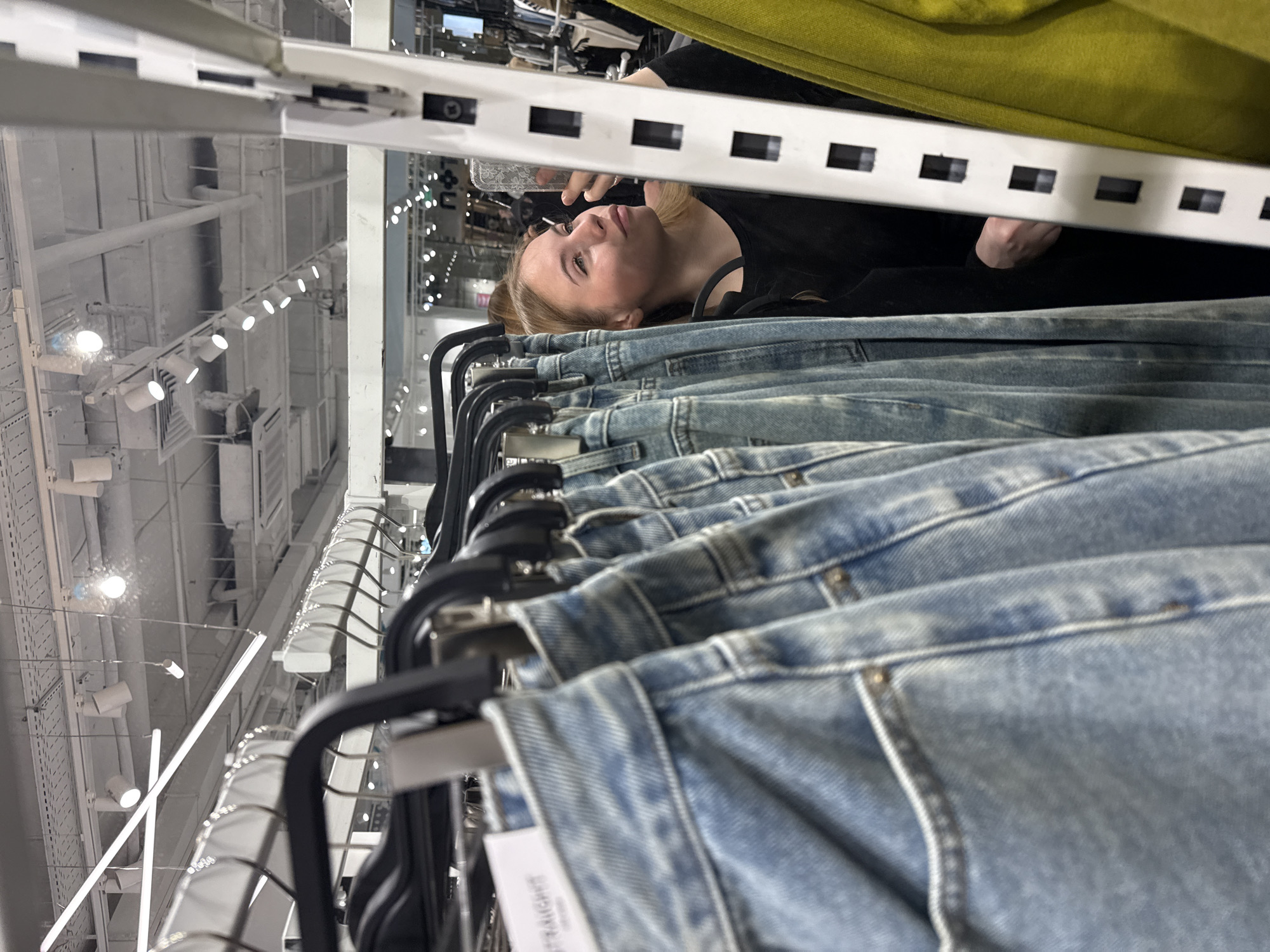 This image depicts a young female inside a retail clothing store, located in Sochi, Russia. In the immediate foreground, a row of light blue denim jeans, identified as "STRAIGHT" fit by a visible tag, hangs on black plastic hangers, rendered out of focus. A young female, with blonde hair and wearing a dark long-sleeved top, is positioned behind these jeans. She is looking into a reflective surface, likely a mirror, and holding a smartphone with a patterned case up to her face, appearing to take a photo or evaluate her reflection. To her right is a white, perforated vertical retail display support, and partially visible beyond it is a bright yellow-green garment. The background shows a brightly lit retail environment with a white industrial-style ceiling, featuring exposed ductwork, electrical conduits, and a variety of lighting fixtures, including long fluorescent tubes, recessed spotlights, and track lighting. Other blurred merchandise displays and signage, including some Cyrillic text, are visible further back within the store.