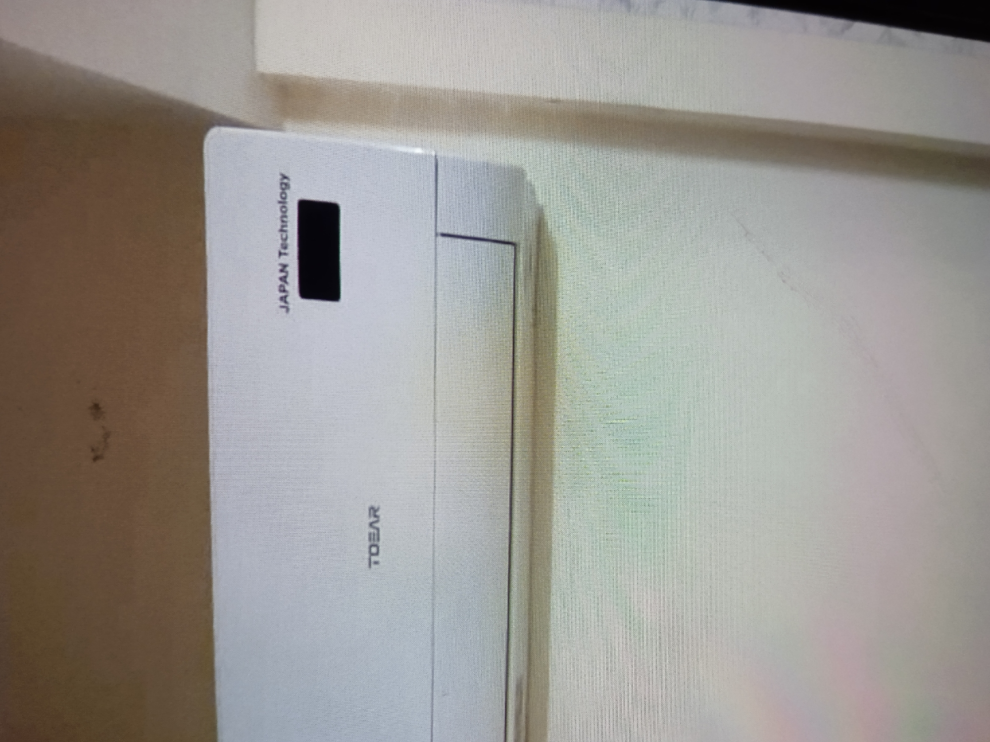The image shows a close-up view of a white air conditioner unit mounted on a wall. The unit is predominantly white with a black rectangular indicator or button on the top left side. Vertical text is printed on the unit, reading "JAPAN Technology" above the black rectangle and "TDEAR" below it. The wall behind the air conditioner appears to be textured and a light beige color. There is a shadow cast on the wall to the right of the unit, suggesting a light source from the left. The overall scene is indoors, and there are no people visible. The lighting is somewhat dim, creating the shadow. The image is angled, with the air conditioner appearing to be on the left side of the frame, and the wall extending to the right. There are some dark spots or stains visible on the wall to the far left of the image, but these are not directly on the air conditioner unit. The image quality is not perfectly sharp, with a slight blurriness and some visible pixelation, suggesting it might be a photograph taken of a screen or at a distance.