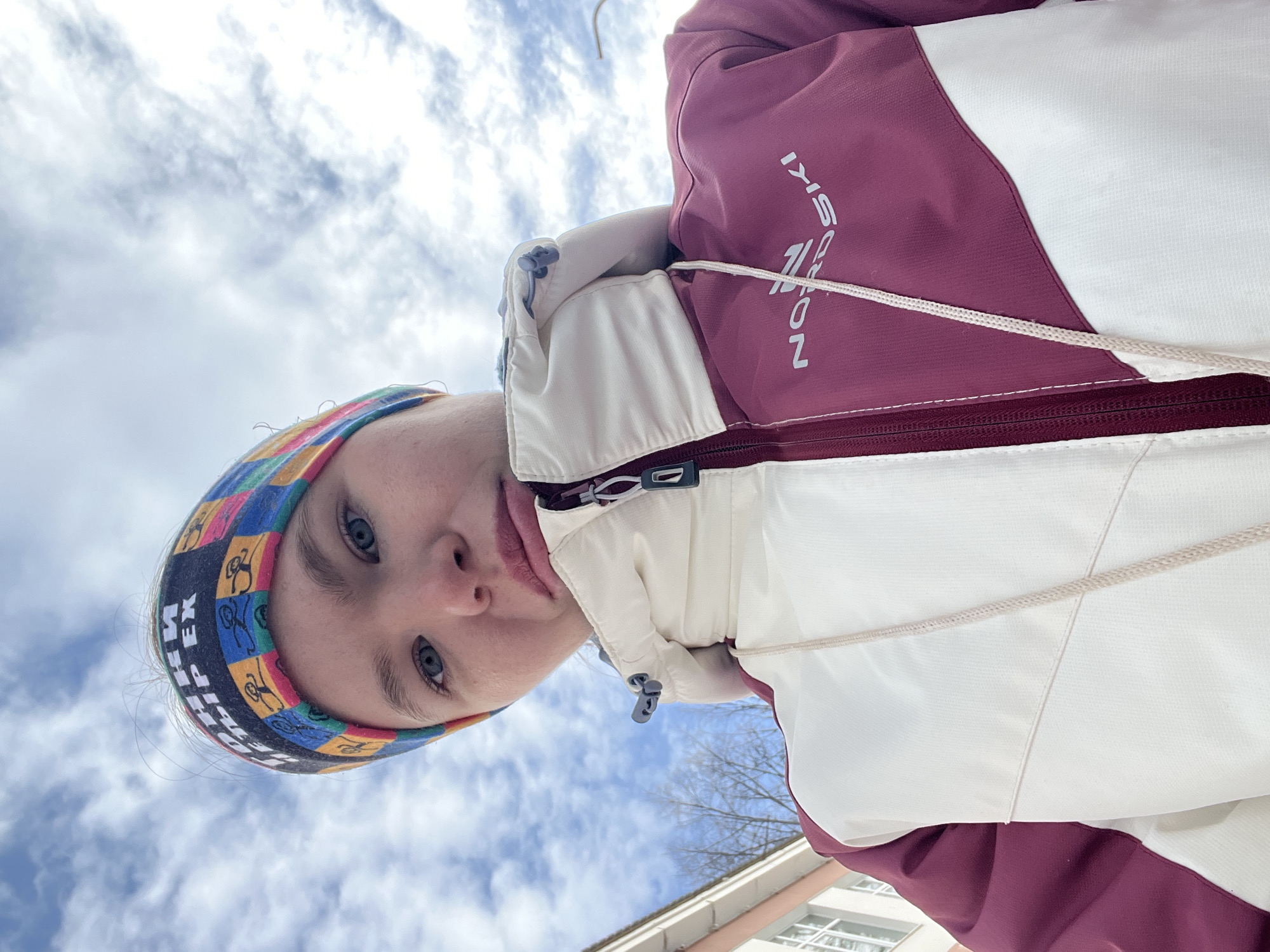 An individual, appearing as a young female, is shown in a low-angle close-up, gazing directly at the camera. She wears a multicolored headband featuring stick figures and Cyrillic text reading "ГОНКИ ПЯТЕР ЕХ". Her eyes are light blue. She is dressed in a winter jacket, half cream-white and half burgundy, with the brand "NORDSKI" visible in white lettering on the burgundy section. A white drawstring hangs from the jacket's collar area. The background consists primarily of a cloudy sky with patches of blue. To the lower left, a segment of a light-colored building with white window frames and several bare trees are visible. The scene is located in the city of Chaikovsky, Russia.