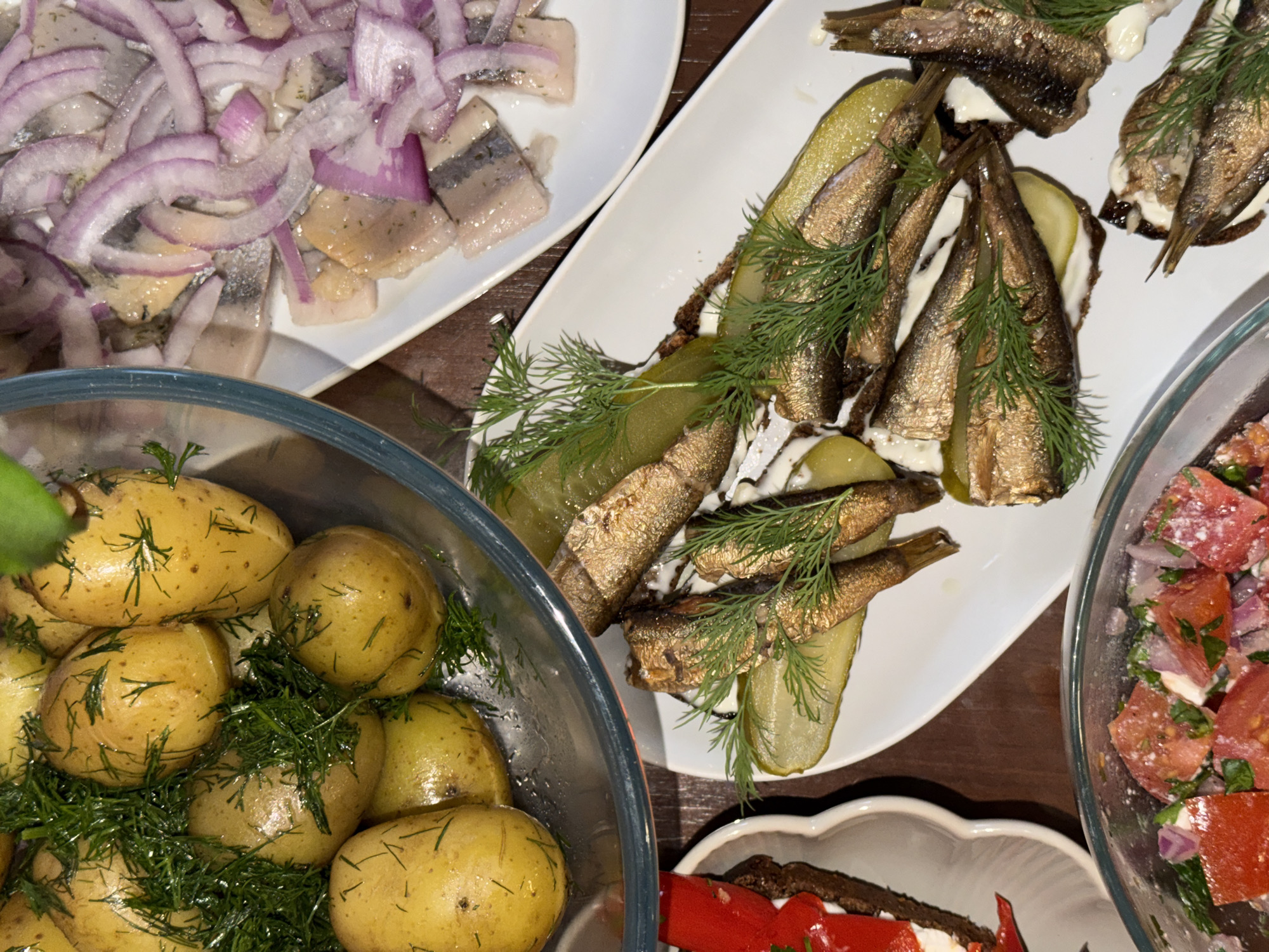 The image shows a tabletop spread of food. There are dishes containing herring with red onion, sprats (small fish) on bread with pickles and dill, small potatoes with dill, and a tomato salad. The scene gives the impression of a traditional Northern or Eastern European meal. The location in the city of Budva, Montenegro, is not evident from the image.