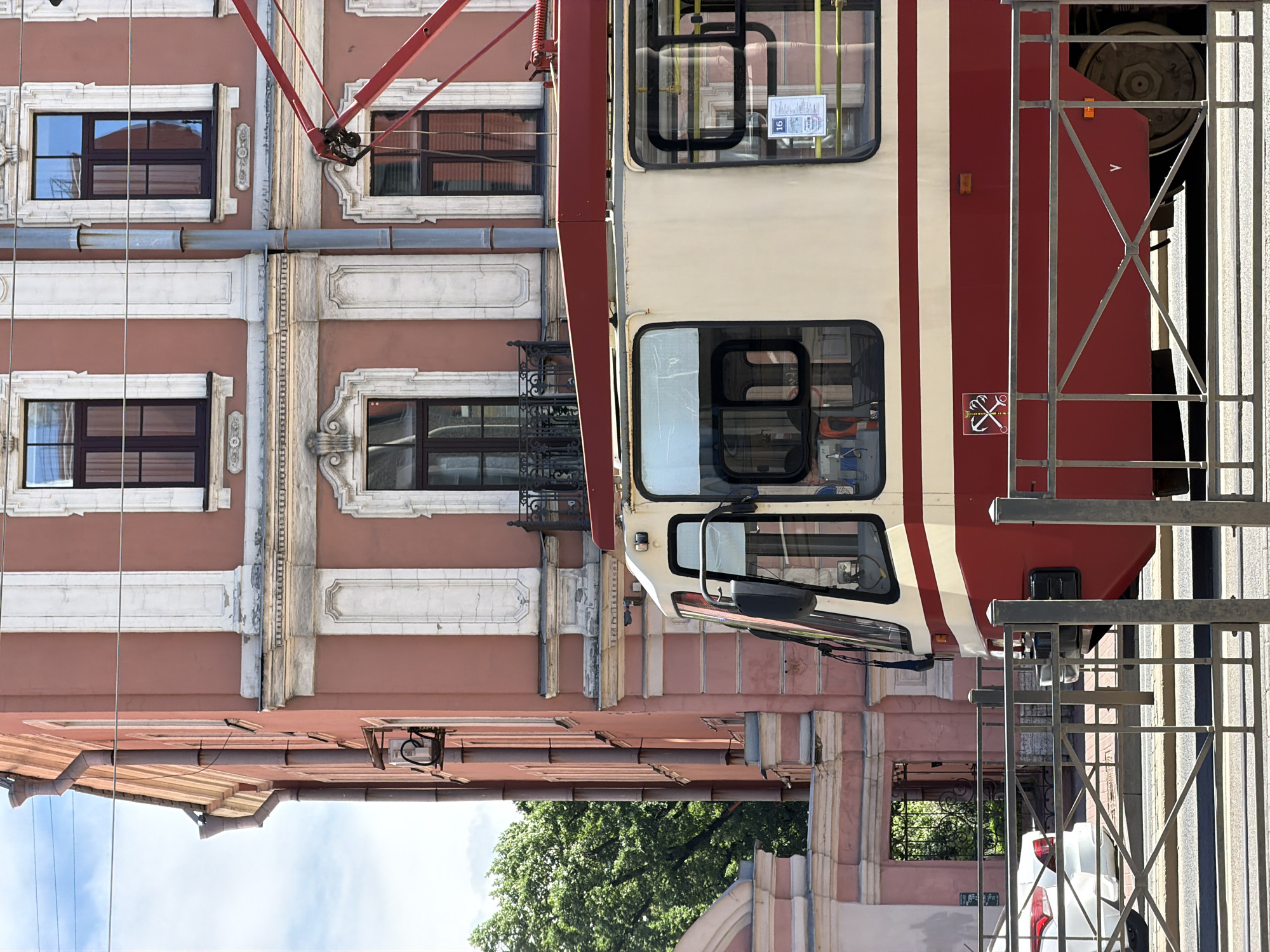 The image depicts a street scene in Saint Petersburg, Russia, under bright daylight conditions. A red and cream-colored tram occupies the midground, partially obscured by a grey metal pedestrian barrier fence in the immediate foreground. The tram's front driver's cabin and a section of its side are visible. A red pantograph extends upwards from the tram's roof. On the red lower panel of the tram, a white rectangular emblem displays the Saint Petersburg coat of arms, featuring two crossed anchors and a scepter with a double-headed eagle. Inside one of the tram windows, a sign with the number "16" is visible.
In the background, a multi-story building with a faded salmon-pink facade stands prominently. This building features numerous white architectural details, including vertical pilasters, ornate window frames, decorative cornices, and horizontal bands separating floors. Dark-framed windows are arranged symmetrically, with one on the second floor featuring a small black wrought-iron balcony. Grey metallic drainpipes run vertically along the building's exterior. An outdoor unit, possibly an air conditioner, is mounted on the left side of the building near a drainpipe.
To the far left, beyond the building and a partially visible white car, dense green tree foliage is present. Further to the left and back, a pink-hued structure with an arched gateway and a dark wrought-iron gate is visible. The sky above is clear blue with sparse white clouds. Strong sunlight creates distinct shadows, indicating a sunny day. No people are explicitly visible in the frame.