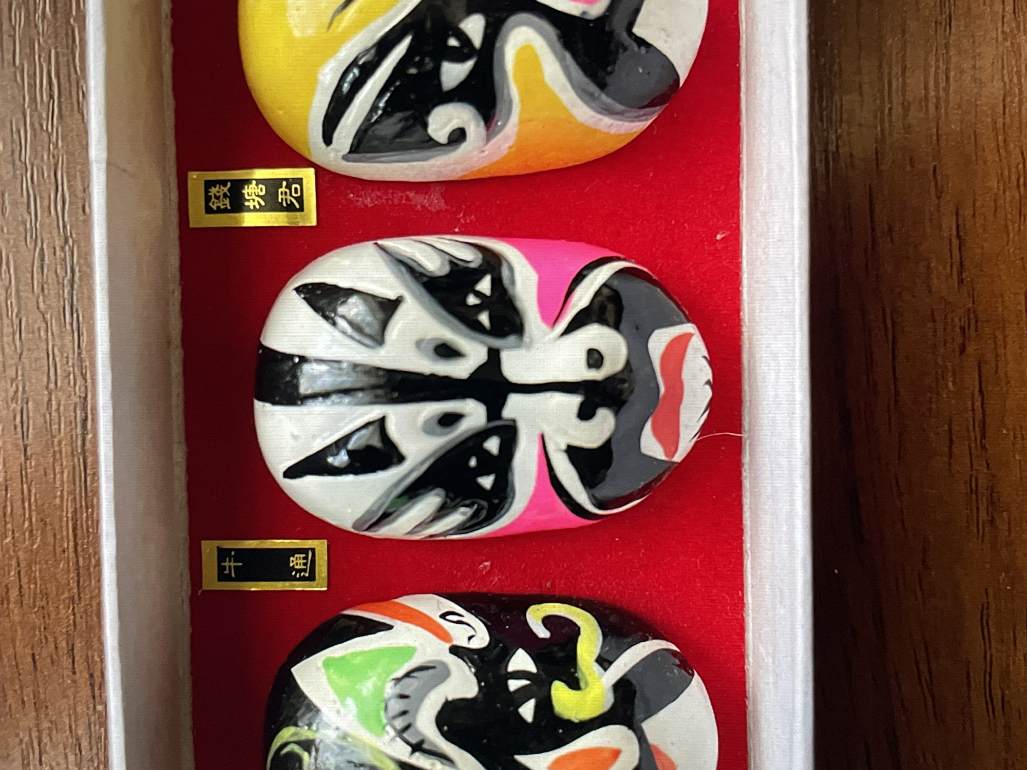 The scene displays three miniature, vibrantly painted Chinese Peking Opera masks arranged side-by-side within a rectangular display box. This scene is located in the city of Saint Petersburg, Russia. The masks rest on a plush red velvet-like lining. The leftmost mask features a predominantly black and white face with green over the left eye, orange on the right cheek, and a yellow stylized mustache. The central mask has a white base, black around the eyes and nose, and prominent hot pink accents on the cheeks with a red lower lip. The rightmost mask is primarily yellow with black patterns defining its features, accented by white lines. Above the masks, two small, vertical golden labels with black Chinese characters are affixed to the red lining; the left label reads "牛通" (Niú Tōng) and the right label reads "錢塘君" (Qián Táng Jūn). The display box features a light-colored, possibly wooden, top edge and a white lower frame, resting on a dark brown surface with visible wood grain.