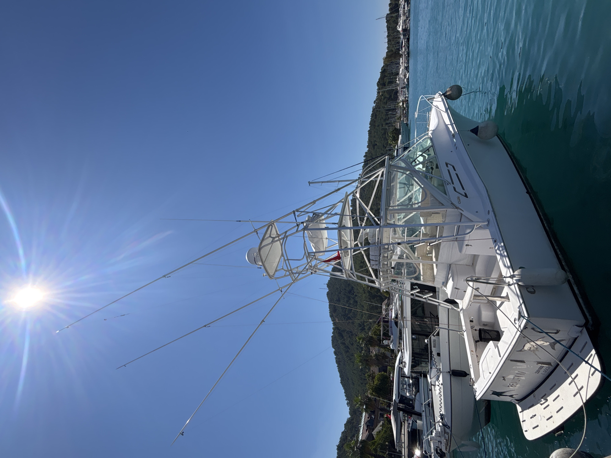 In Fethiye, Turkey, a white sport fishing boat, named "BEAUTY IV," is docked in a clear blue marina. The boat features a prominent silver tower with multiple fishing rod holders extending upwards. Another boat, a white cruiser named "MAVERA," is docked alongside it. In the background, a verdant hillside dotted with buildings and palm trees overlooks the marina, where numerous other boats are moored. The sun shines brightly in a clear blue sky, casting a lens flare. No people are visible on the "BEAUTY IV" or "MAVERA" in this frame.