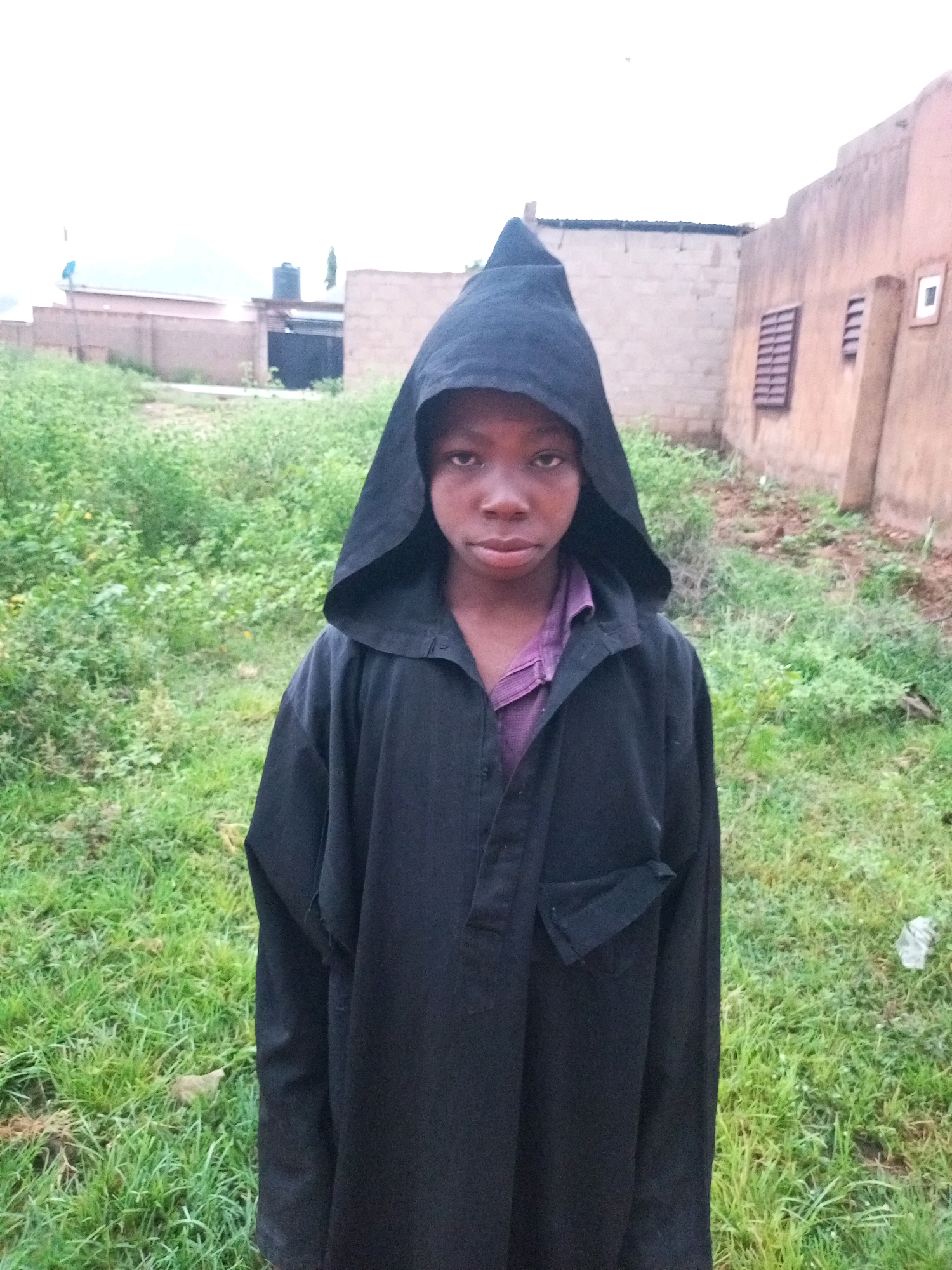 In Tempure, Nigeria, a young boy stands in the foreground, facing the camera. He is wearing a black hooded robe over a purple shirt. The boy's expression is serious, with dark eyes looking directly at the viewer. The robe has a pointed hood and a partial zipper closure at the neck, with a flap pocket visible on the left side.
Behind the boy, the environment consists of a grassy, somewhat overgrown area. In the background, there are structures that appear to be buildings. To the left, a light-colored building with a dark roof is visible, and a black water tank sits atop it. Further back, the sky is overcast. To the right, a light brown building with window-like openings is present. The overall scene suggests an outdoor setting in a residential area.