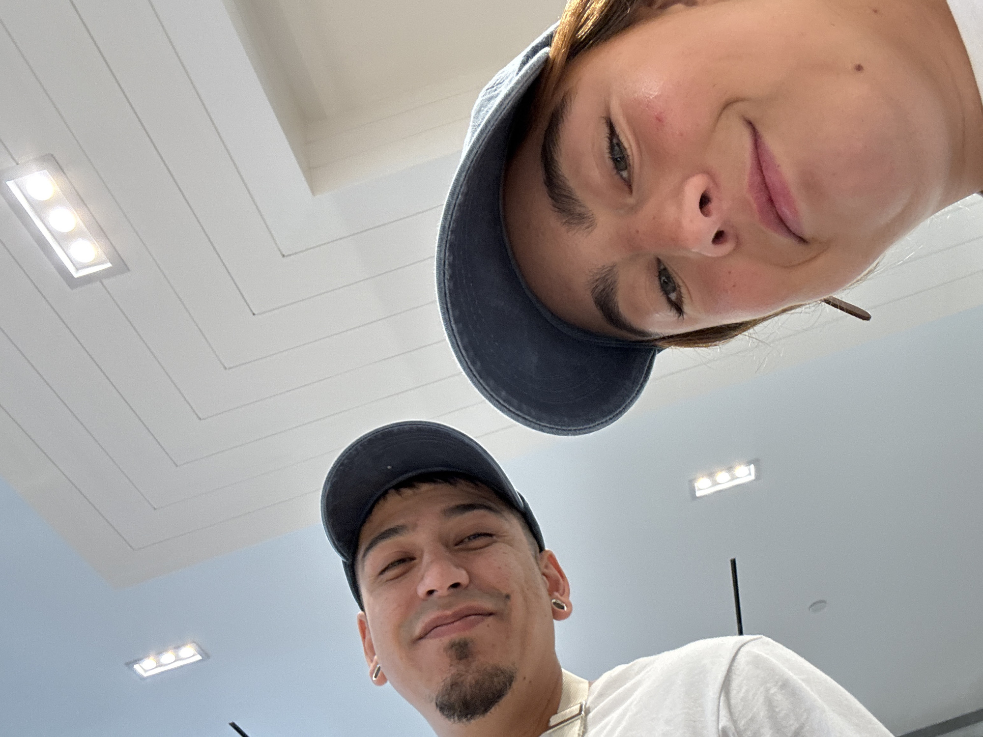 Two individuals are captured in a close-up selfie within an interior space. The ceiling above is visible, featuring a white herringbone pattern with recessed rectangular lighting fixtures. The person at the top of the frame is wearing a blue baseball cap and has light skin, while the individual at the bottom also sports a blue baseball cap and has a goatee. The location may be in the city of West Hollywood, United States of America. No specific actions or interactions are discernible beyond the act of taking a selfie. The scene is dominated by the faces of the two people and the overhead ceiling.