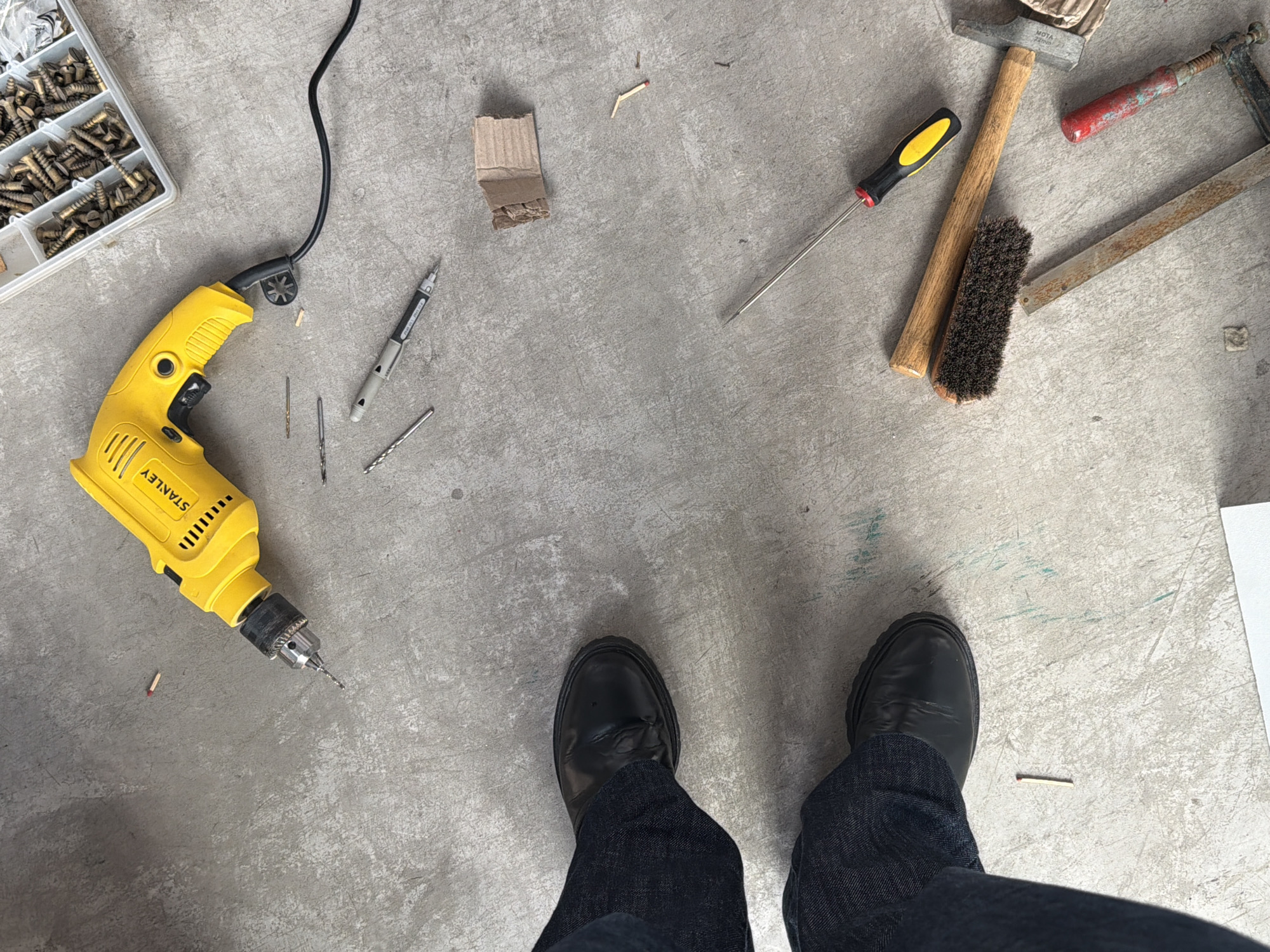 An overhead, medium shot captures a collection of tools and a person's feet on a light gray concrete floor, typical of a workshop or construction area in Buenos Aires, Argentina. The scene suggests active work or preparation for a project.
In the upper left quadrant, a clear plastic organizer box is filled with numerous brass screws. A bright yellow "STANLEY" electric drill, connected by a black power cord, lies adjacent to the screw box, with several drill bits scattered near its chuck. A grey utility knife or multi-bit screwdriver is also visible close to the drill cord.
Towards the upper right, a small cardboard scrap and a few broken matchsticks are present. An assortment of hand tools occupies the right side of the frame: a screwdriver with a yellow and black handle, a hammer with a wooden handle (on which "MOTA - AZOND" is faintly visible), a dark-bristled brush, and a red-handled metal clamp.
At the bottom of the image, the feet of a person wearing black leather shoes and dark blue jeans are seen, standing on the concrete floor. Another matchstick lies just in front of the right shoe. The lighting is uniform and bright, suggesting an indoor daytime setting.