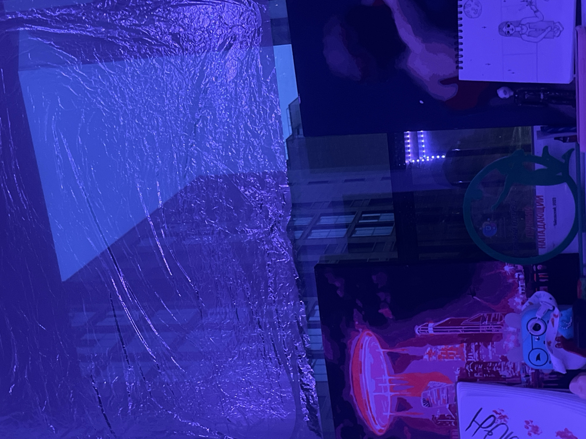 The image shows a scene indoors, illuminated by a blue light. On the left side, there's a shiny, wrinkled material covering an indiscernible surface. Through a gap between this material and a dark, flat object, a glimpse of a building can be seen, suggesting the location is a window or reflective surface overlooking an urban area.
To the right, there are several objects stacked on a surface. There's a spiral-bound notebook with a drawing of a figure. Below that, there is a green circular object and a print with artwork depicting fantastical buildings or space objects. On top of the prints, there's a blue object that may be a toy or gadget, and a white card.
Without additional information, the specific location in Saint Petersburg cannot be determined.