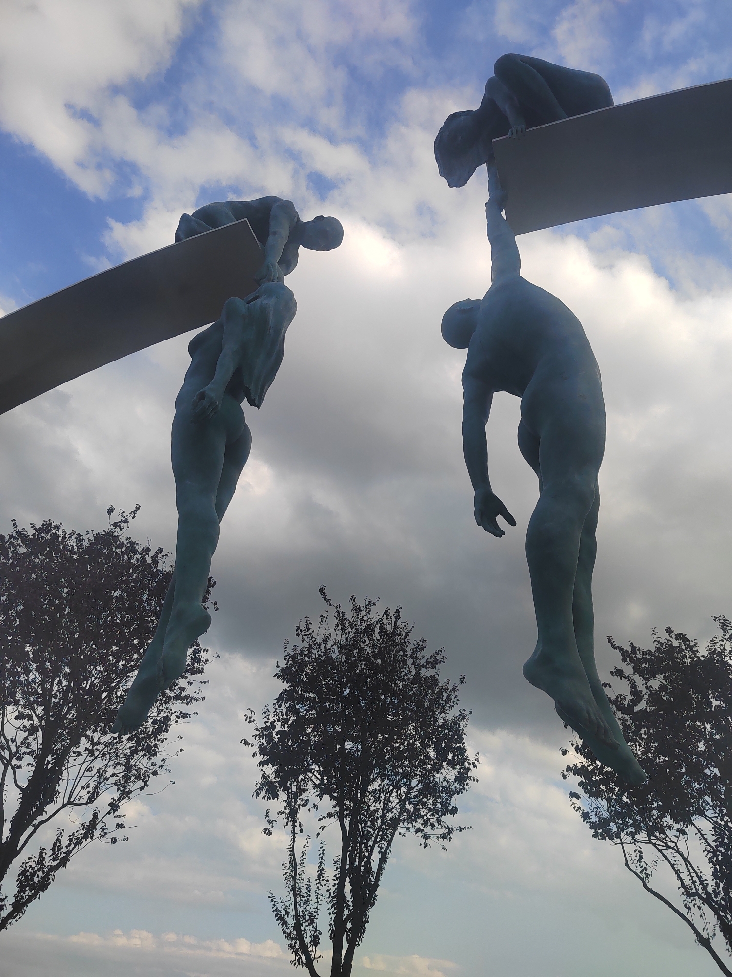 This is a low-angle, full-shot photograph of a sculpture in Vityazevo, Russia, depicting three figures in a dynamic pose. Two figures are suspended from a horizontal beam, with one figure gripping the beam with its hands and the other hanging below, reaching up to clasp the hand of the figure above. A third figure is partially visible, crouched on the beam, appearing to be in the act of reaching down. The sculptures are made of a teal-colored material, and their musculature is clearly defined. The background consists of a cloudy sky and the silhouetted branches of two trees on either side of the frame.