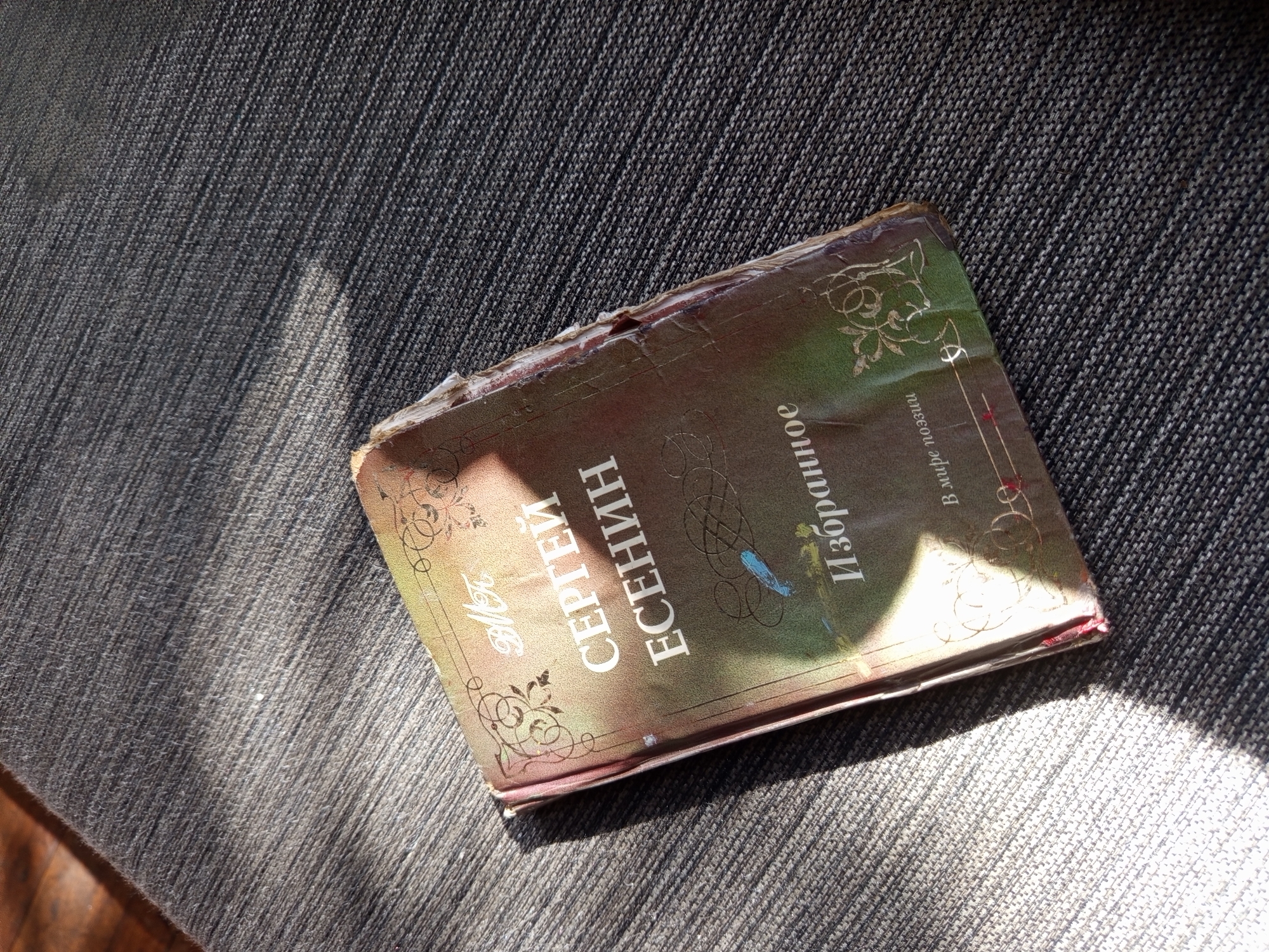 A worn copy of a book titled "Sergei Yesenin Selected" in Russian, with a brownish-green cover and decorative patterns, rests on a textured grey surface, likely fabric. Sunlight casts shadows across the scene. The book is aged, showing wear on the edges of its cover. The location is not identifiable.