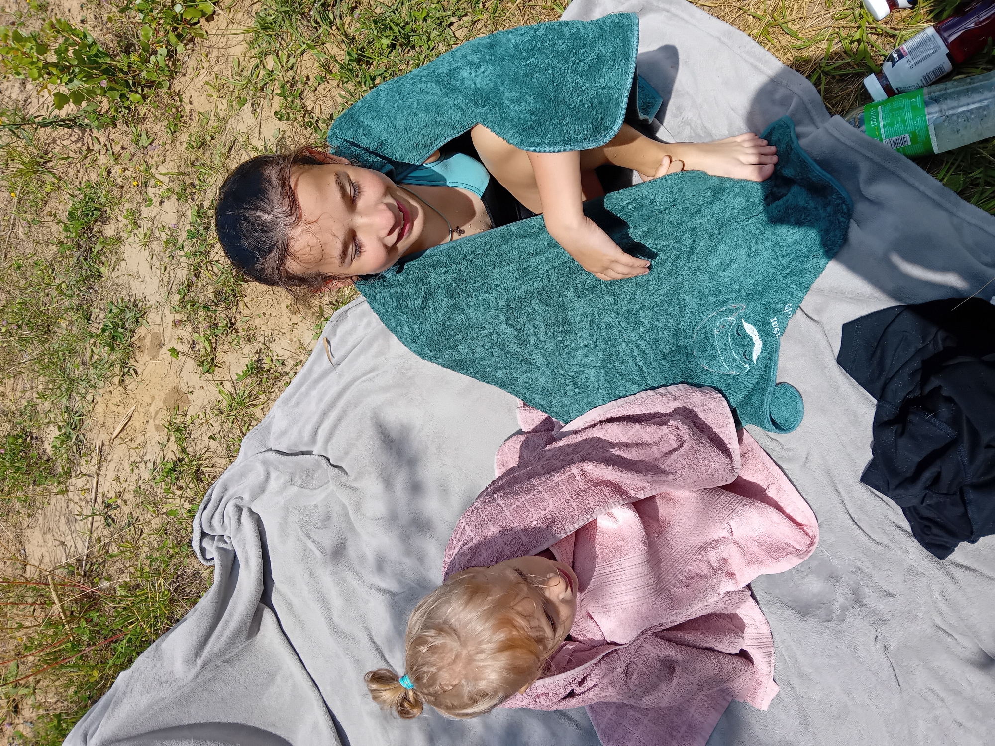 Two young females, one older and one younger, are sitting on a gray blanket outdoors on a sunny day. The older female, on the right, is wrapped in a teal towel and wearing a blue and black swimsuit. She has her eyes closed and is smiling. The younger female, on the left, is wrapped in a pink towel and has her head bowed, looking down. They are surrounded by dry grass and dirt. In the lower right corner, there are two plastic bottles, one clear and one dark purple, and a black piece of clothing. The overall scene suggests a relaxed, outdoor activity, possibly after swimming. The location is Chuhuiv, Ukraine.
