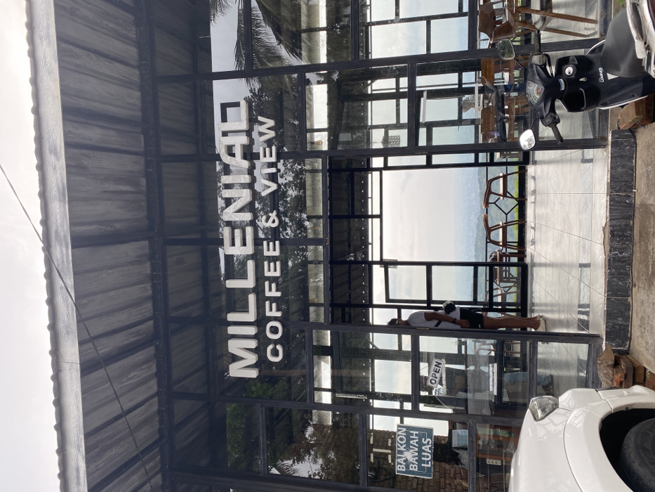 The image shows the entrance of a coffee shop called "Milleniad Coffee & View" located at the coordinates -7.855, 110.473 in Kecamatan Piyungan, Indonesia. The building is made of black metal framing with large glass windows. An "OPEN" sign is hung on the door. A man wearing a white shirt and black shorts is standing in the doorway. He is facing the viewer. A black scooter is parked in front of the doorway. The scooter's handlebar is visible. The interior of the coffee shop is visible through the glass doors. There is a small table and chairs placed in front of the door on the left side of the image. The exterior of the shop is on the right side of the image. It is a large, open area with a view of distant mountains. The sky is cloudy and overcast. There is a sign that says "BALKON BAWAH LUAS" which translates to "lower balcony spacious" hanging on the left side of the door frame. The sign is written in Indonesian. The exterior wall of the building is made of brick.