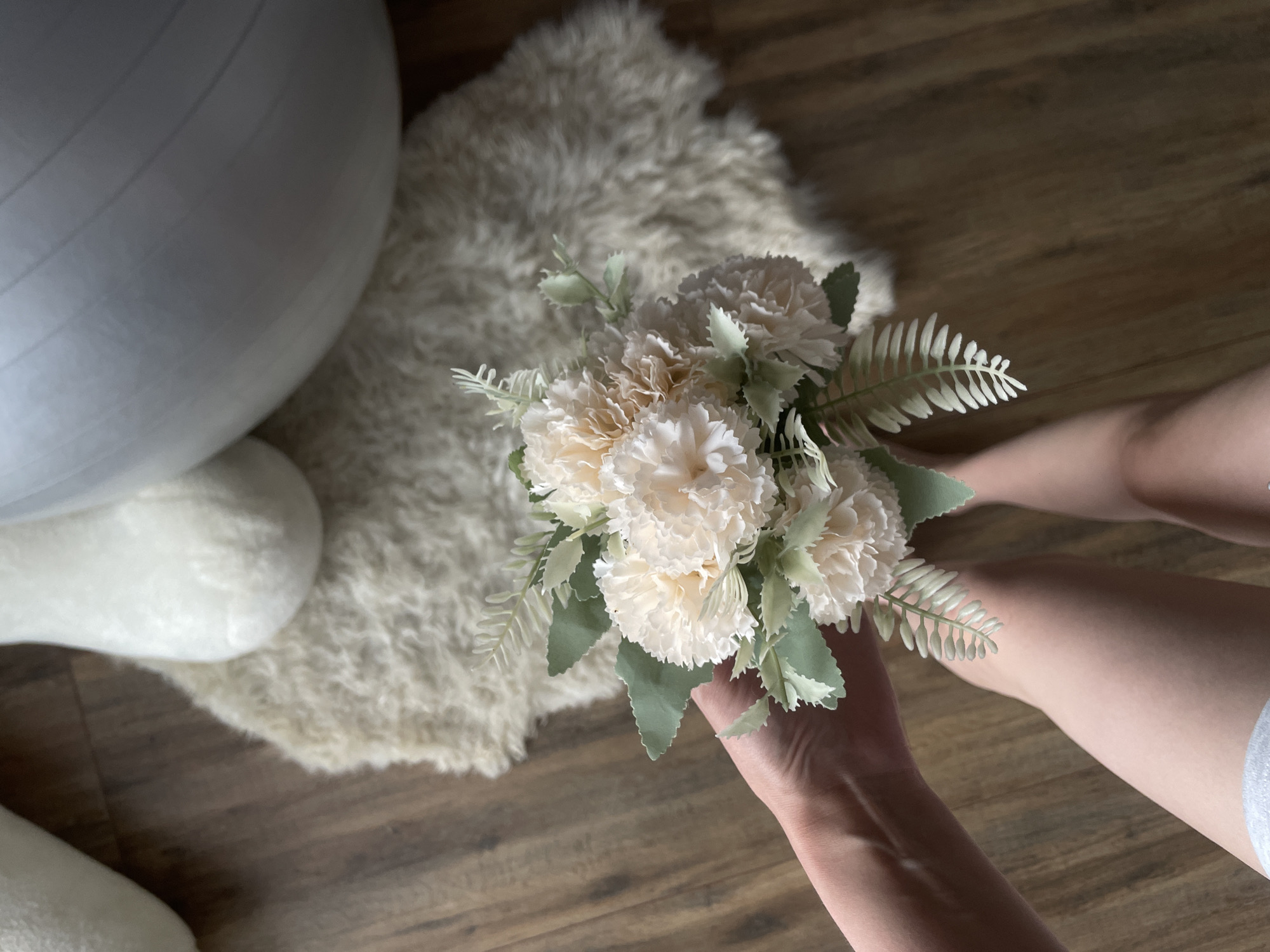 An overhead, high-angle shot depicts an indoor scene in Podshivalovo, Russia. A person's right hand is prominently featured in the lower right, holding a small bouquet of artificial flowers. The bouquet consists of several light beige or off-white blossoms, resembling carnations, mixed with various light green artificial foliage, including some distinctly fern-like leaves.
The person's lower legs and feet, partially covered by light-colored clothing or socks, are visible on the right side of the frame, suggesting a seated position on the floor or a low surface. In the upper left quadrant, a large, light grey exercise ball or similar rounded object is partially visible. Underneath the ball and extending towards the center of the image is a fluffy, cream-colored rug or sheepskin, adding a soft texture to the setting. The background consists of a warm-toned wooden floor with visible planks and grain. The lighting is soft and ambient, characteristic of an indoor environment. No text is visible in the image.