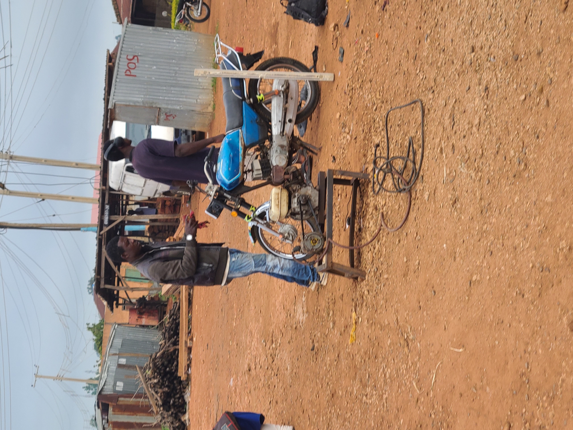The image depicts an outdoor scene in Barikin Sabon Gidan Kanat, Nigeria. In the foreground, a blue and silver motorcycle is parked on reddish-brown dirt. A portable generator is situated in front of the motorcycle, connected by wires to something out of frame. Two men are present. One man, wearing a dark jacket over a white shirt and blue jeans, stands to the left of the motorcycle, appearing to be working with something red in his hands. The other man, wearing a dark purple shirt and black headphones, is partially visible behind the motorcycle, facing away from the camera. In the background, various structures are visible, including a corrugated metal shed with "POS" written in red letters, a wooden stall, piles of logs, and a white van. Electrical poles and overhead wires are prominent in the sky. The overall environment suggests a semi-urban or market area, possibly a workshop or service point.