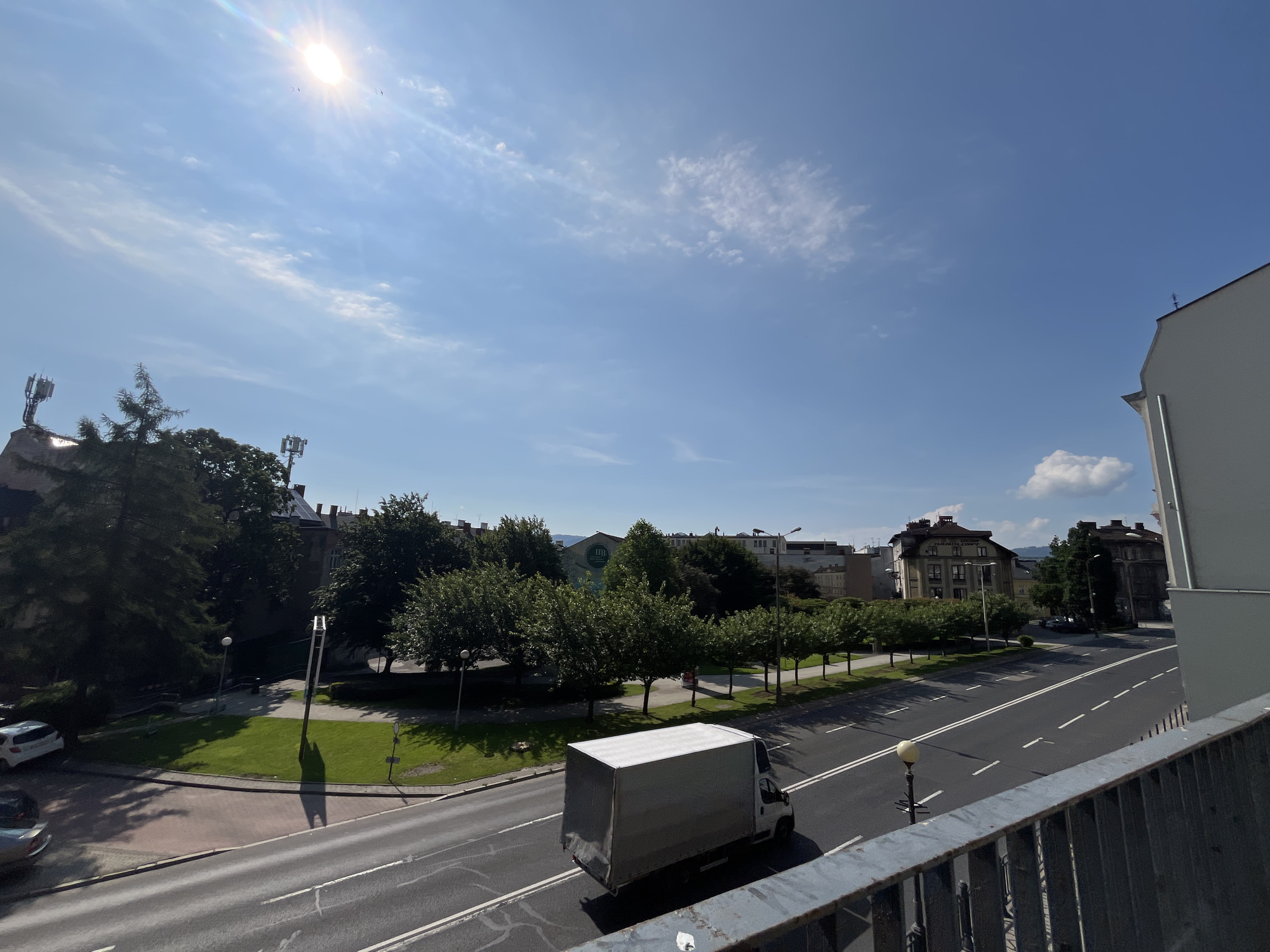An outdoor, eye-level, wide shot captures a sunny day in an urban environment. The bright sun illuminates a clear blue sky with scattered white clouds. In the foreground, a weathered metal railing overlooks a city street. A white box truck travels down the road, casting a shadow on the asphalt. To the left of the truck, a strip of green grass is bordered by a curved red path. Mature trees with lush green foliage line the street, casting dappled shadows. In the background, buildings of varying architectural styles are visible. Two cellular antennas are prominent atop a dark building on the far left. The scene conveys a sense of everyday city life under a clear sky.