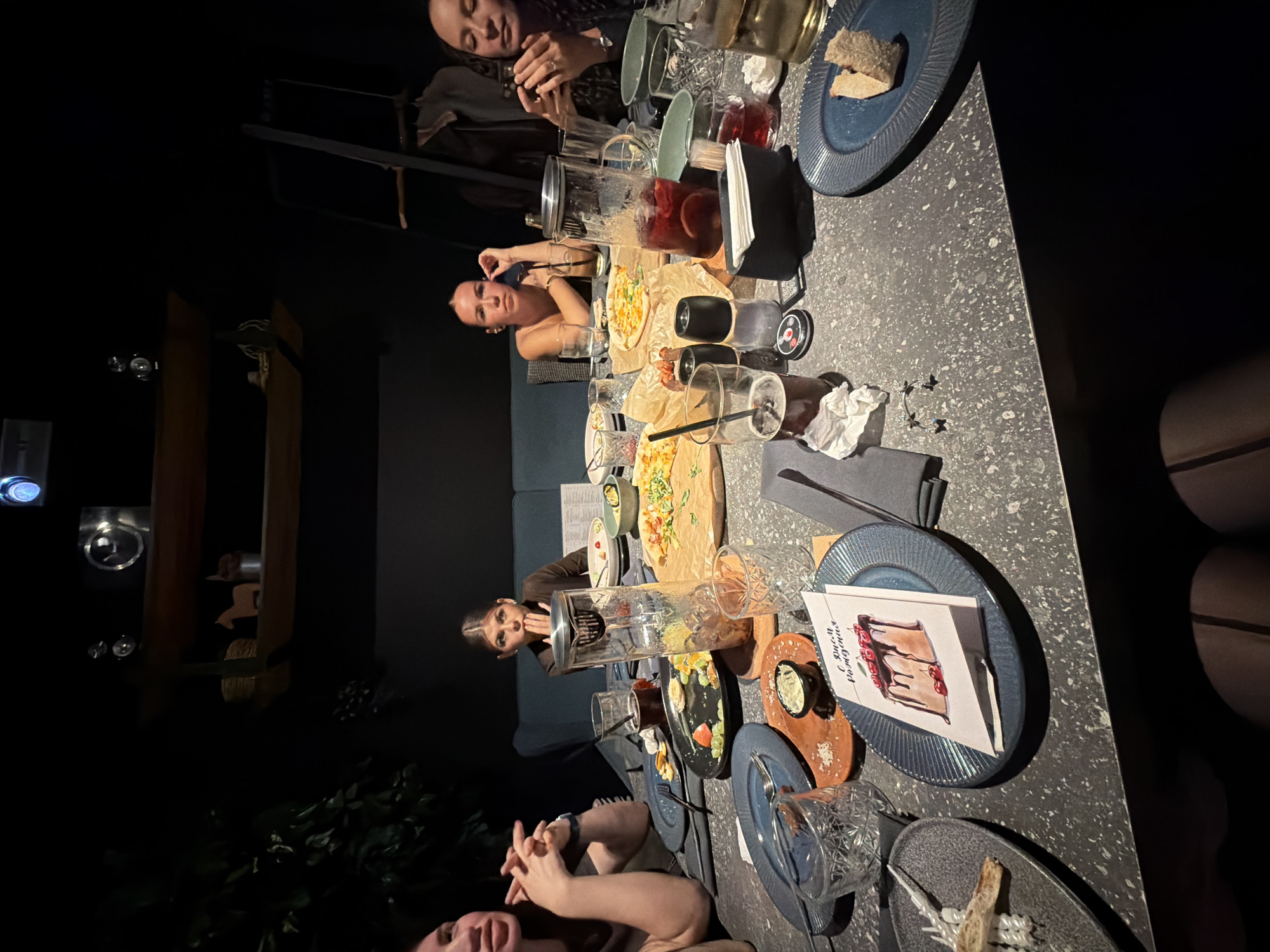 The image depicts an evening gathering of approximately five to six individuals, predominantly women, seated around a large, dark grey speckled table in what appears to be a dimly lit restaurant or bar in Chaikovsky, Russia. The group is engaged in dining and socializing, with several individuals looking towards the camera or conversing, conveying a relaxed and celebratory atmosphere.
The table is well-stocked with food and drinks. Visible dishes include multiple small pizzas or flatbreads served on wooden boards lined with paper, a salad or mixed appetizer in a bowl, and bread slices on plates. Beverages include a large pitcher of a red drink, possibly sangria, along with various glasses, some containing drinks with ice and straws, and others empty.
Other table items include blue and dark grey plates, clear glasses, black and white crumpled napkins, and small black butterfly-shaped decorations. A key detail is a white greeting card placed on a blue plate, adorned with an illustration of a chocolate cake with cherries. The legible text on this card, "С днем рождения!", which translates to "Happy Birthday!", indicates that the occasion is a birthday celebration.
The lighting is dim, suggesting a nighttime or evening setting, with reflections from glassware and light sources in the dark background. The scene captures an intimate and enjoyable birthday dinner or get-together.