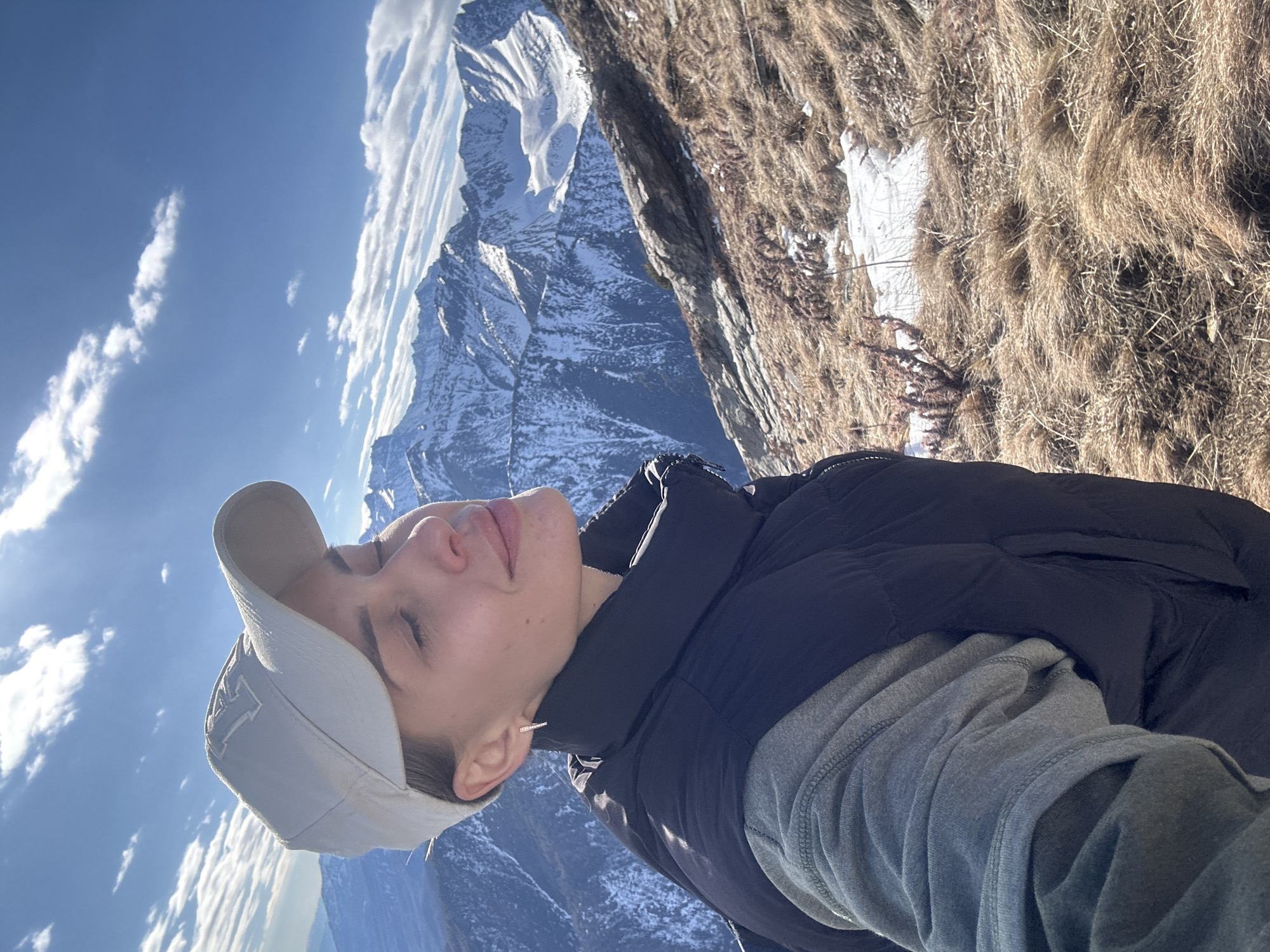 A person wearing a light gray baseball cap, a gray long-sleeved shirt, and a black puffer vest is positioned on a mountain ridge, likely in a selfie perspective. The person's eyes are closed, and they are facing towards a partly cloudy sky with bright sunlight. Behind the person, there is a steep, rocky, mountainous terrain with patches of dry brown grass and snow. In the distance, snow-capped mountains are visible under a clear blue sky. The overall scene suggests a location high in the mountains, with signs of late autumn or early spring weather. The photo was taken in Arshan, Russia.