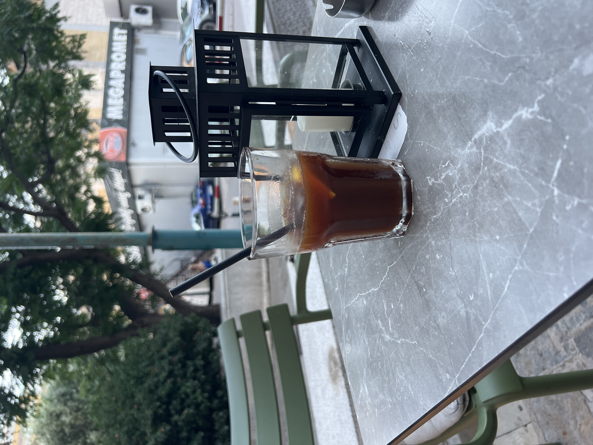 An iced coffee sits on a marble-topped outdoor cafe table, next to a black metal lantern with a white candle inside. The glass is condensation-covered and contains a dark beverage with ice and a straw. In the background, a green metal chair is visible, along with trees and buildings. A sign that reads "MEGAPROMET" is also visible. The scene appears to be outdoors during the daytime, possibly in a European city. The overall mood is relaxed and casual.