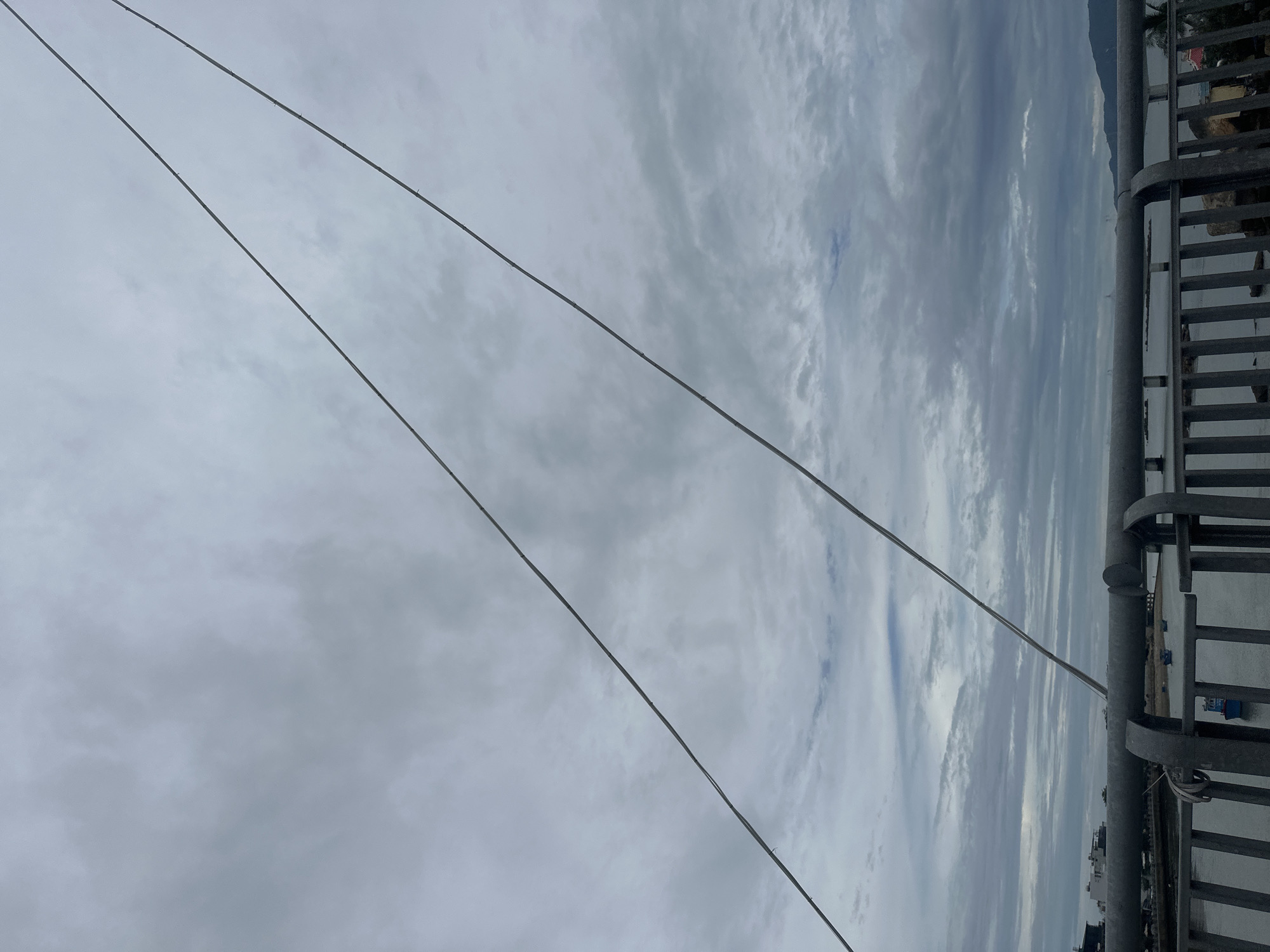 The photograph, taken in Bắc Nha Trang Ward, Vietnam, depicts an overcast sky with layered clouds. In the lower portion of the frame, there are two metal wires running diagonally from the upper left to the lower right corner. On the right side of the image is a metal railing. Beyond the railing, there is a blurred view of what appears to be buildings and potentially a body of water.
