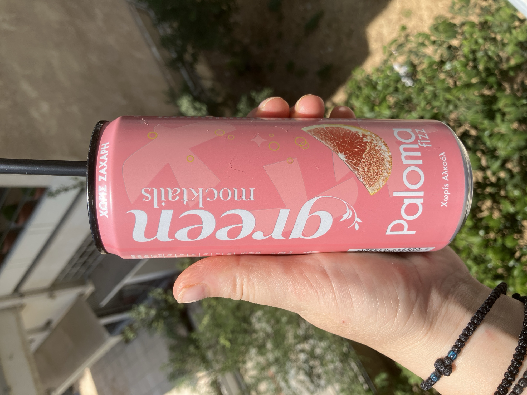 A person's hand holds a pink, slim can of "green mocktails" with "Paloma fizz" written in large white letters and "χωρίς ζάχαρη" (without sugar) at the top. The can has a grapefruit slice graphic and a black straw. The background shows a slightly out-of-focus building and green foliage, suggesting an outdoor urban setting. The scene could be from Paleo Faliro, Greece, given the Greek text on the can.
