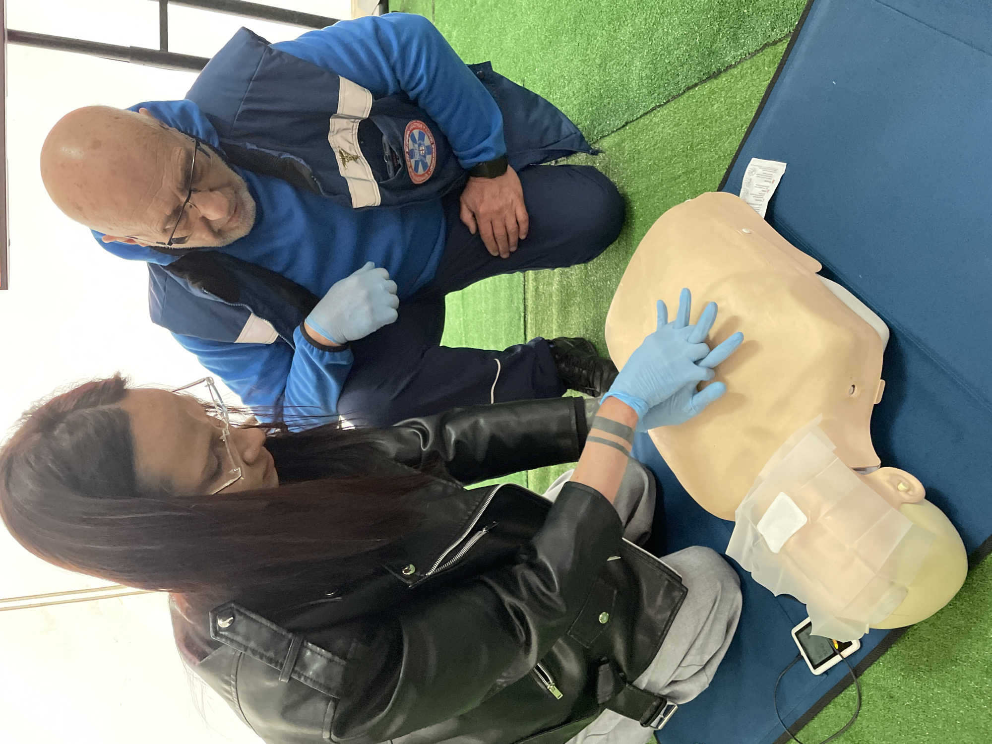 Two individuals are engaged in cardiopulmonary resuscitation (CPR) training within an indoor facility in Yekaterinburg, Russia. A younger adult female, with long dark hair and wearing clear-framed glasses, a black leather jacket, and grey trousers, is kneeling on the left. She is performing chest compressions on a light-skinned, beige CPR training manikin, using both hands interlocked over the manikin's sternum. She wears light blue disposable gloves. An older adult male, bald with a grey beard and wearing glasses, a blue hooded sweatshirt, and a dark blue sleeveless vest with reflective white strips, kneels to the right of the female, observing her actions. He also wears a light blue disposable glove on his left hand. The male's vest displays a silver Caduceus emblem on the right chest and a circular blue, white, and red emblem with a Star of Life-like symbol on the left chest pocket. The manikin is placed on a rectangular blue mat, which rests on green artificial turf flooring. A small white digital device, connected by a black wire to the manikin's head, provides feedback during the training. The background consists of plain white, canvas-like walls, suggesting a temporary structure or training tent.