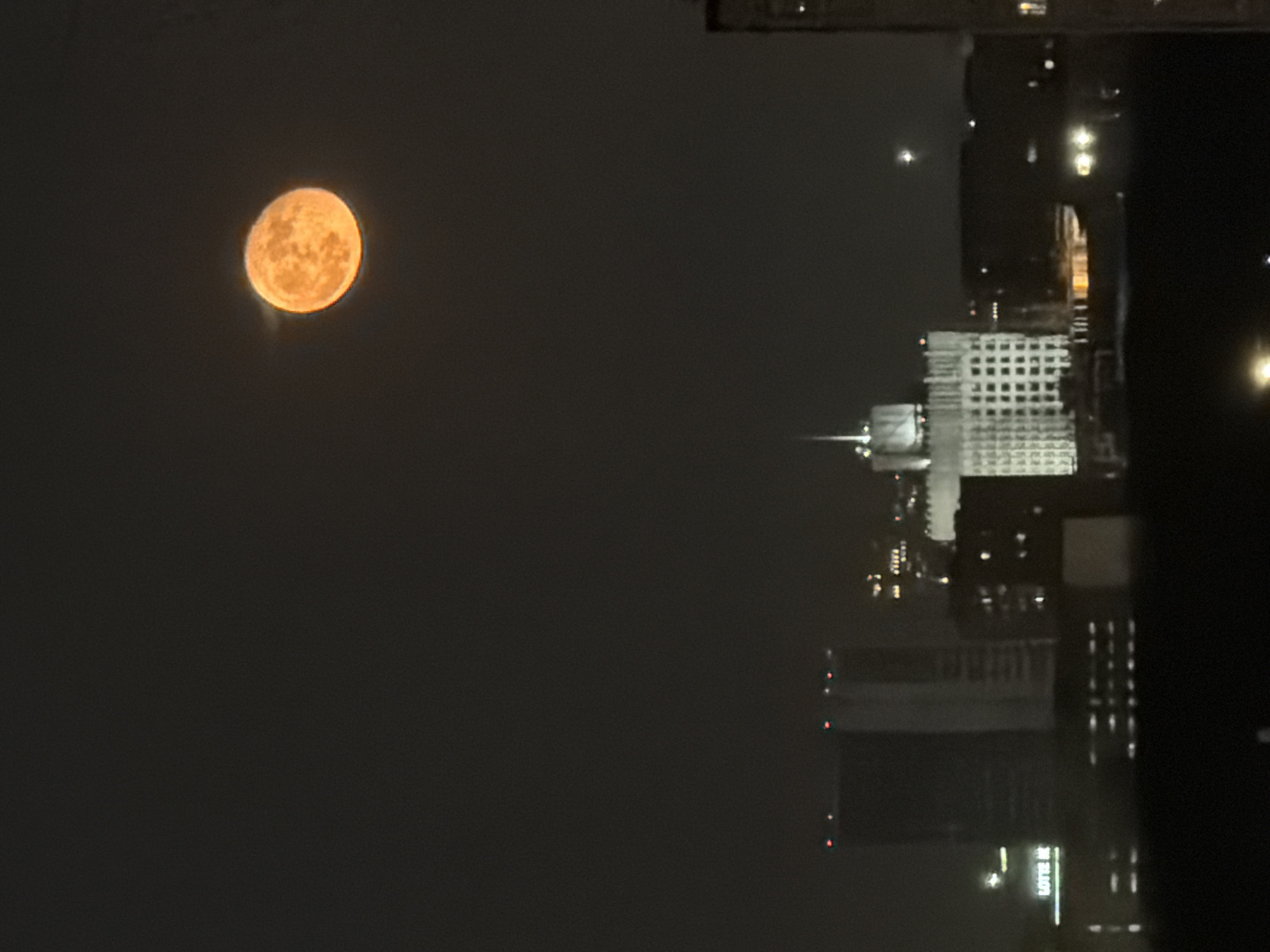 The image depicts a night scene in Khimki, Russia. In the left portion of the image, a large, orange moon is visible in the dark sky. To the right, the cityscape is evident, including buildings of varying heights and illuminated windows. Notably, the reflection of the city lights and buildings is captured, possibly indicating the presence of a body of water or a reflective surface in the foreground. A prominent building with a grid-like pattern of windows is noticeable, along with other structures of modern architectural design. "LOTTE" can be seen at the base of the structures reflecting off the water. Overall, the image captures the contrast between the natural element of the moon and the urban landscape of Khimki, Russia.