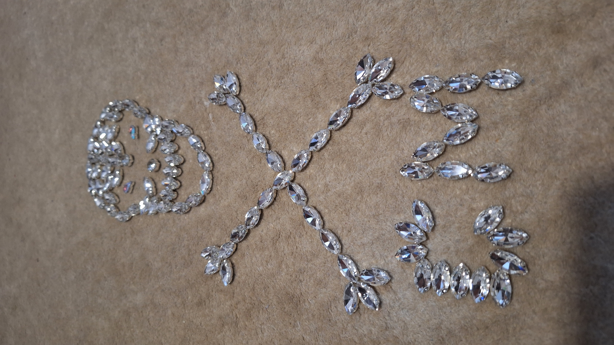 The image captures a close-up view of clear, faceted gems arranged on a textured, light brown fabric background, possibly suede or felt. The primary subjects are three distinct shapes formed by these sparkling stones, which spell out the letters "OXO."
On the far left, a large, intricate circular design represents the first "O." This shape features an outer ring composed of marquise-cut gems, with several smaller, varied-cut gems scattered inside the circle, creating a decorative, almost floral or clock-like pattern. In the center of the frame, a stylized "X" is formed by two intersecting lines of marquise-cut gems. Each arm of the "X" extends outwards, terminating in a cluster of three marquise gems, giving it a star-like or snowflake-like quality. To the right, the final "O" is represented by a simpler ring of marquise-cut gems.
The gems are highly reflective, catching the light and creating bright glints across their facets. The background fabric provides a soft, neutral contrast to the brilliance of the stones. No specific activity is depicted other than the deliberate arrangement of the gems. The scene is an indoor, static display, with no visible time of day, weather, or location cues other than the neutral background. The visible text clearly spells "OXO" using the arranged gems.