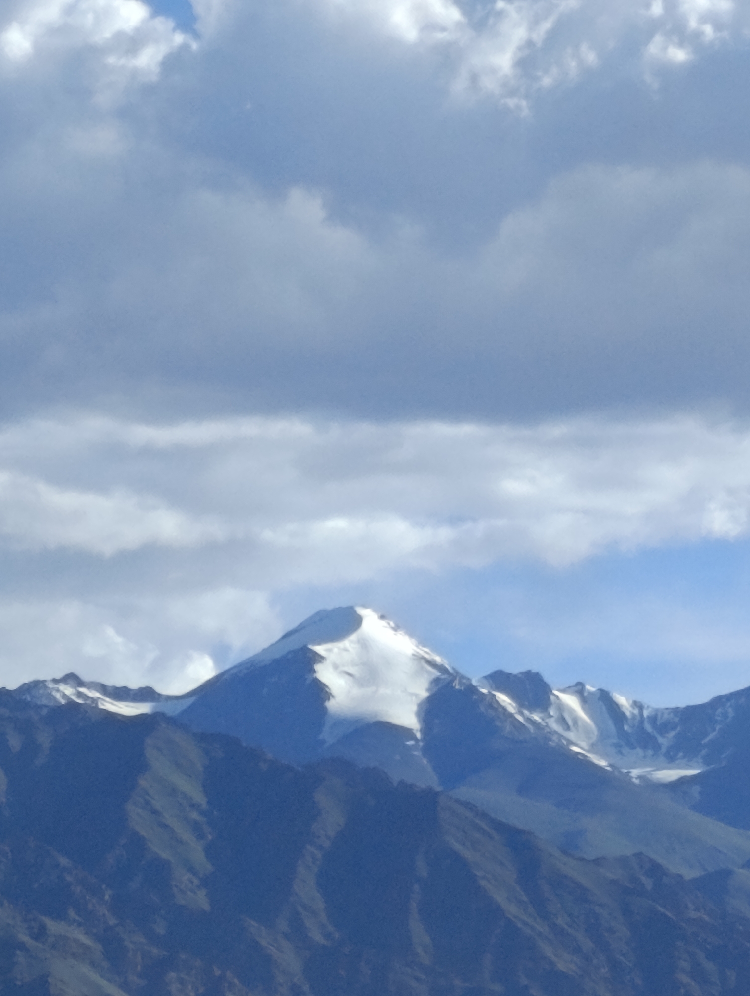 The image shows a mountain range under a cloudy sky. The peak of the main mountain is capped with snow. The lower slopes of the mountains are dark and rugged, with some patches of green visible. The sky is a mix of blue and white clouds, with some darker, heavier clouds towards the top. The scene appears to be a natural landscape with no visible man-made structures or people. The image quality is somewhat soft, suggesting it was taken from a distance or with a lower resolution camera. This scene is characteristic of the mountainous terrain surrounding Leh, India, a region known for its high altitude and dramatic landscapes.