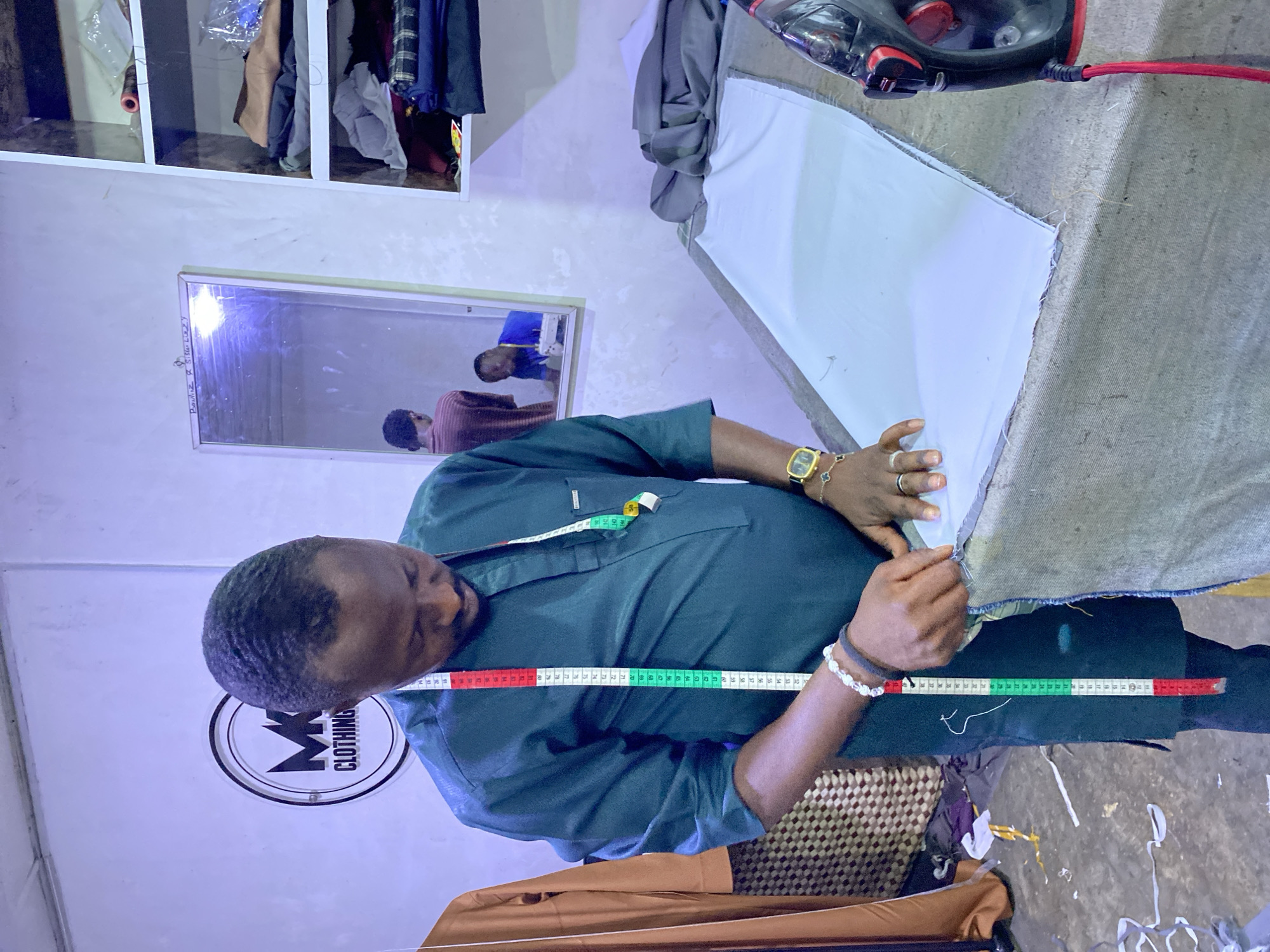 This horizontal image captures a man, presumably a tailor, engaged in his work within what appears to be a workshop or clothing store in Jos, Nigeria. The man, positioned over an ironing or cutting table, is wearing a dark green long-sleeved shirt with a colorful measuring tape draped around his neck. He has a gold watch on his left wrist, and rings and bracelets on his right hand.
He is focused on a piece of white fabric laid out on a large table, which is covered with a textured grey fabric. His hands are visible, smoothing or adjusting the white material. To the right on the table, a red and black iron is present.
The background to the left features white shelving units stocked with various fabrics and clothing items. Below the shelves, a rectangular mirror reflects the room, showing the man's reflection along with partial reflections of two other individuals, one in a brown/red shirt and another in a blue shirt. Text written on the mirror includes "BAWU..." and "SISTAR LAZY".
Behind the man, a circular logo on the wall displays the text "M CLOTHING". The floor, partially visible at the bottom, transitions from a textured mat to a concrete surface littered with fabric scraps and threads. The scene depicts a diligent work process in a functional, well-used indoor setting, likely under artificial lighting.