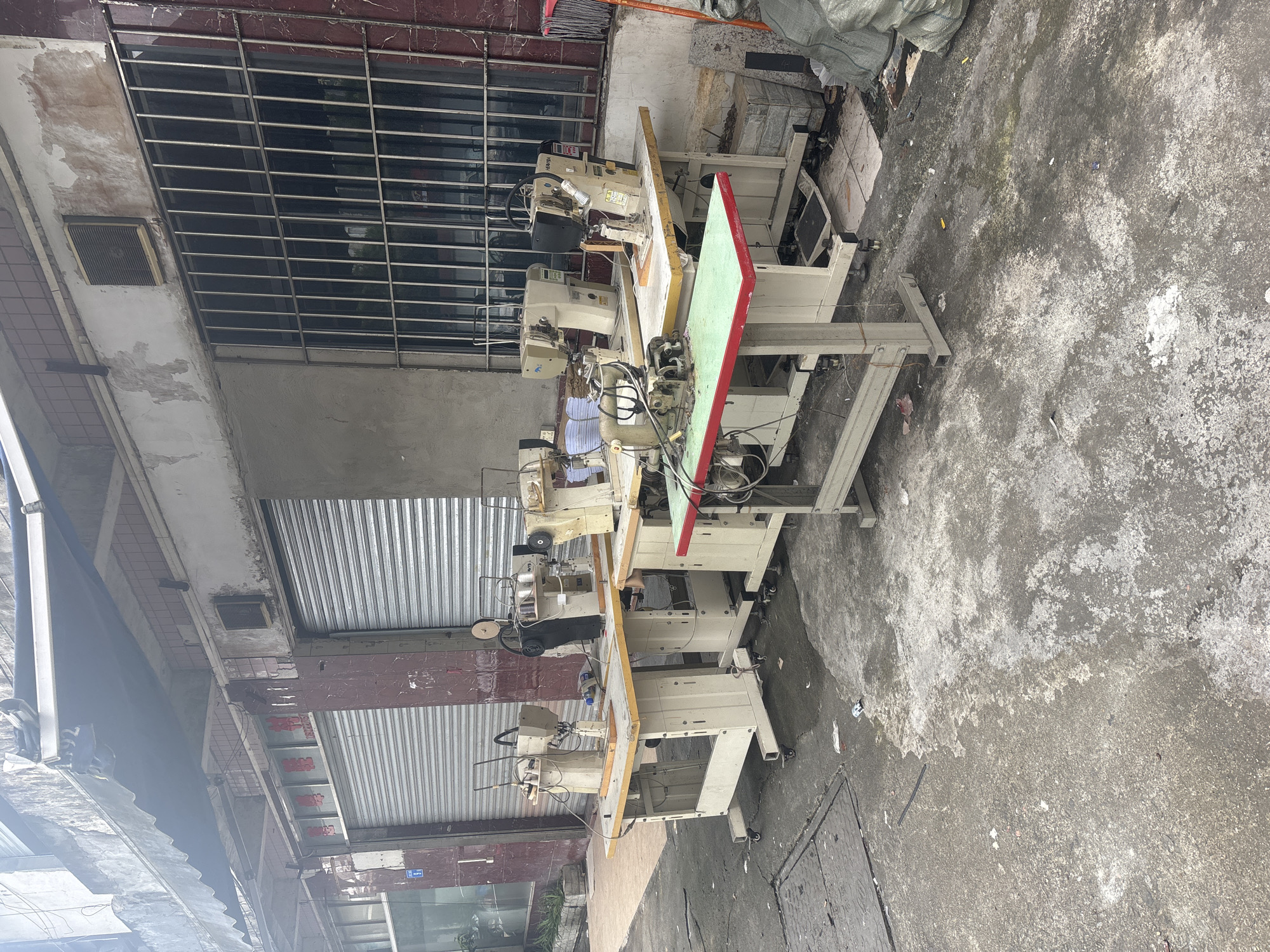 In Nanhai District, China, an outdoor area in front of a commercial building is depicted. The scene features several industrial sewing machines set up on tables. The building's facade includes closed metal roll-up doors and a window with metal bars. The ground is paved with concrete and shows signs of wear and some scattered debris. A large gray bag is visible on the right side of the image.