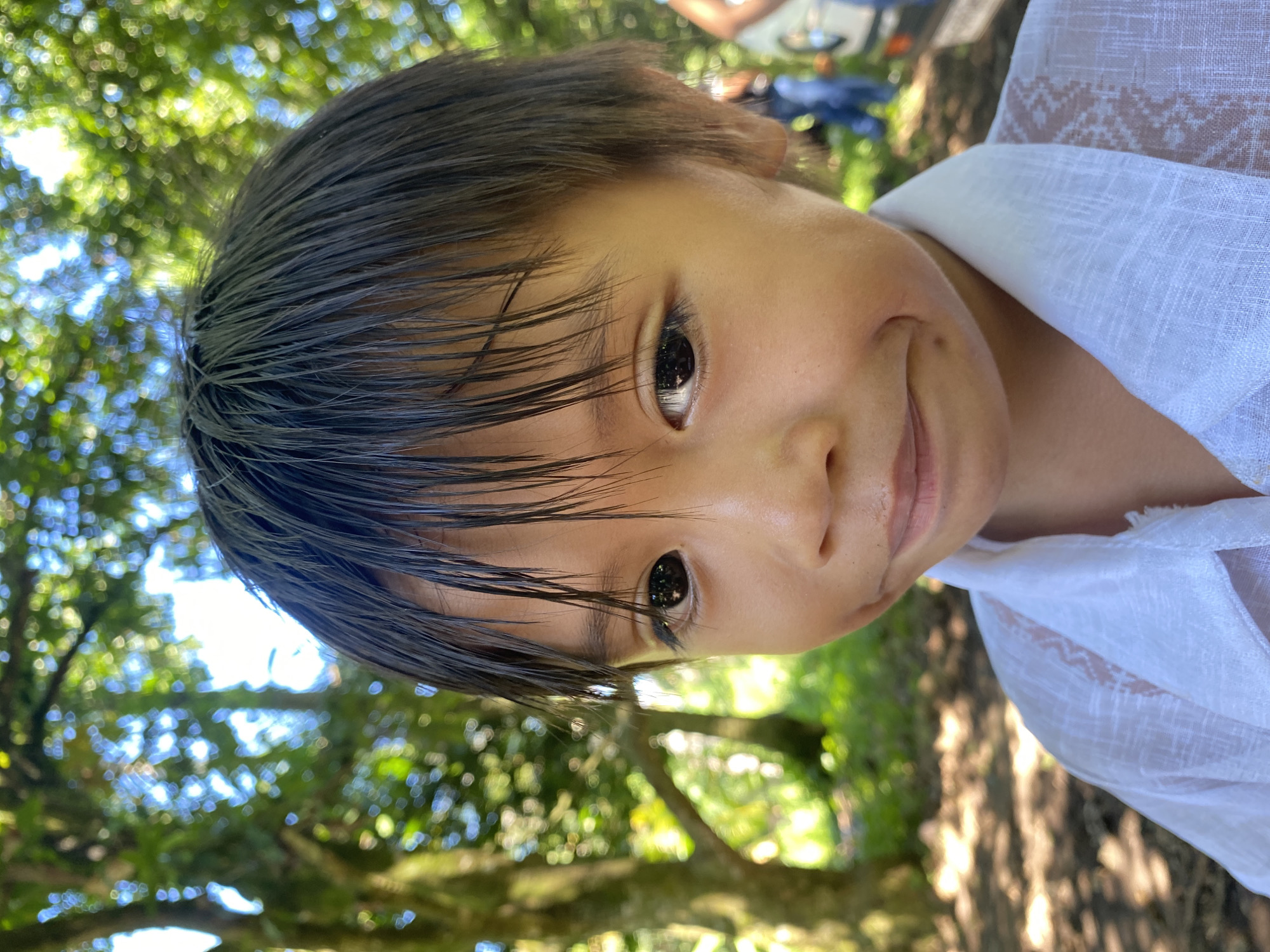 A close-up, eye-level shot captures the upper body of a young Filipino child with dark hair and brown eyes, looking slightly to the right with a shy smile. The child is wearing a white collared shirt with intricate patterns on the shoulders. The background is softly blurred, showing lush green trees and dappled sunlight filtering through the leaves, suggesting an outdoor setting in a park or natural area. Two individuals and a vehicle are partially visible in the far background. The scene evokes a sense of a warm, sunny day, possibly in a park within Davao City, Philippines, known for its abundant greenery and tropical climate.