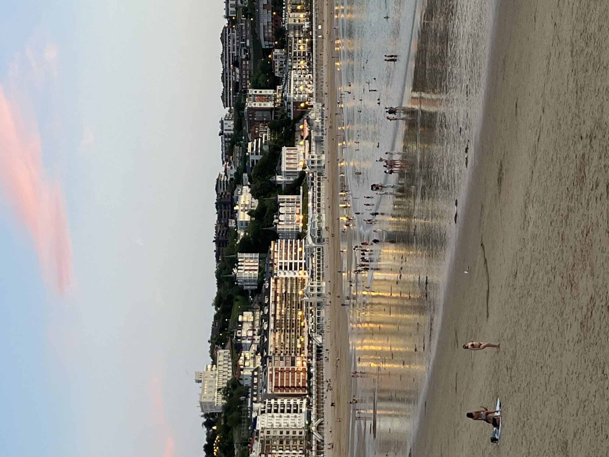 This is a photograph of La Concha beach in San Sebastián, Spain, during twilight. The sky is a pale blue with streaks of pink from the setting sun. The city skyline is visible in the background, with a prominent white building atop a hill, likely a hotel or historical site, followed by rows of multi-story buildings with illuminated windows. The beach is populated by numerous people, some walking along the shore, others wading in the shallow water. The wet sand reflects the lights from the city buildings, creating a shimmering effect. In the foreground, two women are visible on the sand, one sitting and the other standing, both in swimwear. The overall scene depicts a lively evening at a popular urban beach.