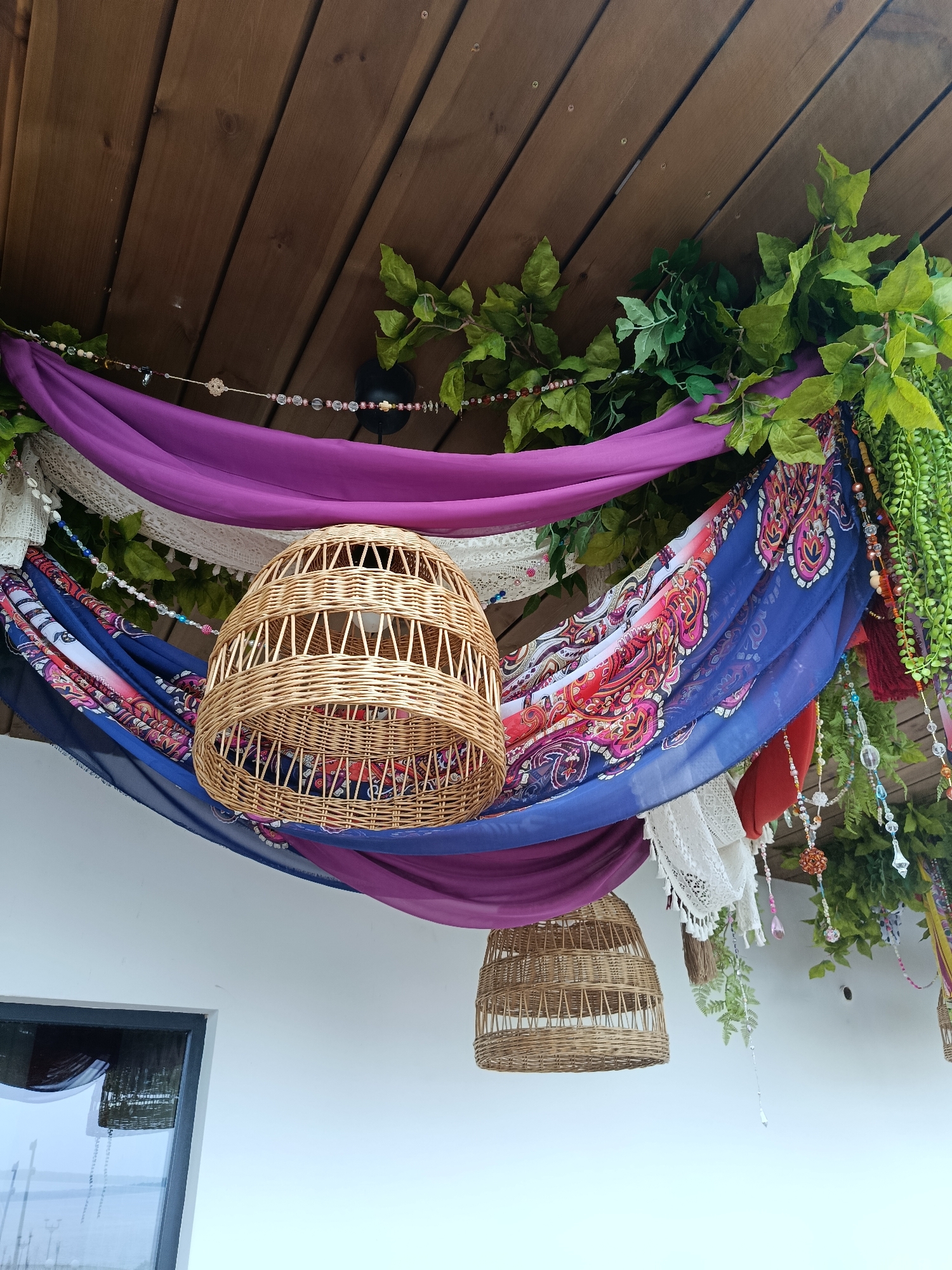 This is a low-angle, outdoor, eye-level shot of a decorated ceiling or awning area. The ceiling is constructed of wooden planks, running diagonally from the top left to the bottom right of the frame. Suspended from the ceiling is a bohemian-style decoration featuring draped fabrics in shades of purple, blue with a paisley pattern, and white lace. Interspersed with the fabrics are strings of colorful beads and tassels, as well as artificial green foliage. Two woven wicker lampshades are hanging from the ceiling, one larger and closer to the camera, the other smaller and further back. Below the decorated ceiling is a white wall, and on the left side of the frame, a dark-framed window with a reflection of what appears to be a body of water and some lampposts is visible. The overall impression is one of a whimsical, outdoor seating or decorative area. There is no indication of the specific location within Saratov, Russia.