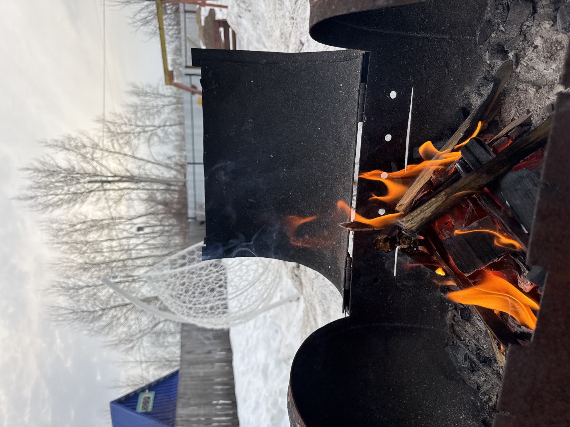 The scene depicts an outdoor setting in Chaikovsky, Russia, during what appears to be winter or early spring. In the immediate foreground, a dark, cylindrical metal barbecue grill contains a vibrant fire with bright orange flames consuming wooden pieces and emitting light smoke. Charred wood and ash are visible at the bottom of the grill. The grill's hinged lid is open and propped upwards.
Beyond the grill, the ground is covered in undisturbed snow. To the left, a white, open-weave egg-shaped hanging chair stands on the snowy ground. A dark wooden fence runs horizontally across the mid-ground. Behind this fence, on the left, a building with a steep, pointed blue roof and a small, rectangular window with a light-colored frame is partially visible. In the center-right background, a cluster of tall, deciduous trees with bare branches extends upwards against a pale, overcast sky. Faint overhead utility lines are visible against the sky above the trees. To the far right, a yellow-colored metal structure, possibly part of playground equipment, is partially discernible above the fence, also on snow-covered ground. No individuals are present in the image.