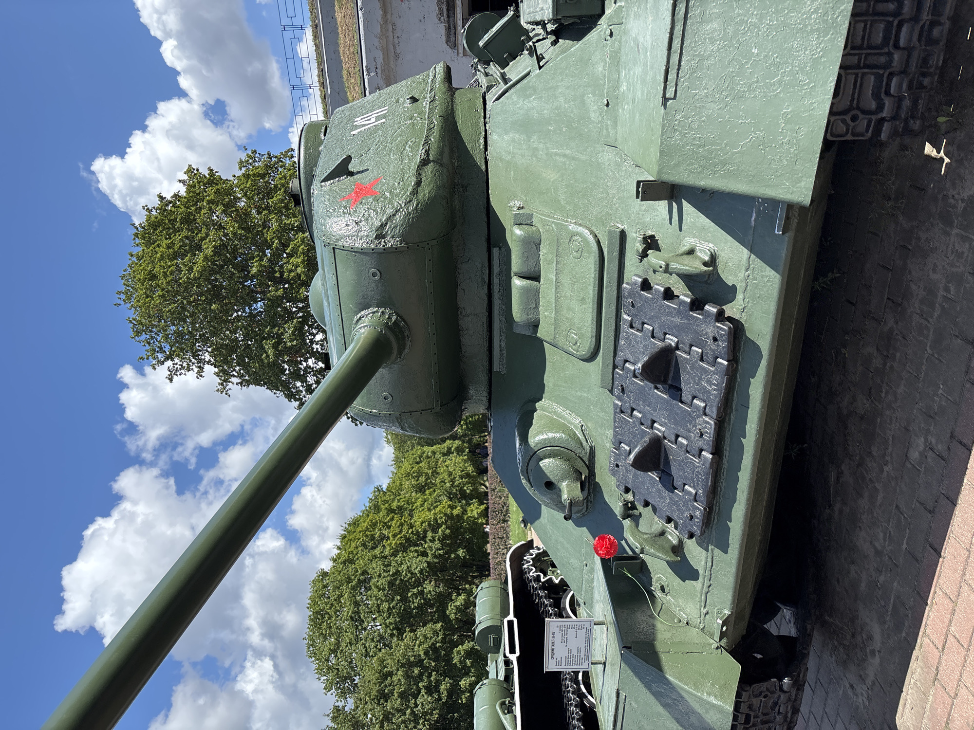 The image shows a close-up, low-angle view of a green Soviet T-34-85 tank displayed outdoors. The tank is positioned in a courtyard with a red brick pavement in the foreground. Behind the tank, there are trees and a blue sky with white clouds. The tank has a red star emblem on its turret and the number "141" is visible. A small red flower is placed on the front of the tank. Another tank, possibly a different model, is visible in the background to the left, with a plaque providing information about it. The scene appears to be a memorial or a museum exhibit. Based on the presence of a T-34-85 tank, it is highly probable that this image was taken at the Museum of Brest Fortress Defense in Brest, Belarus, as this museum features several tanks and military equipment related to World War II.