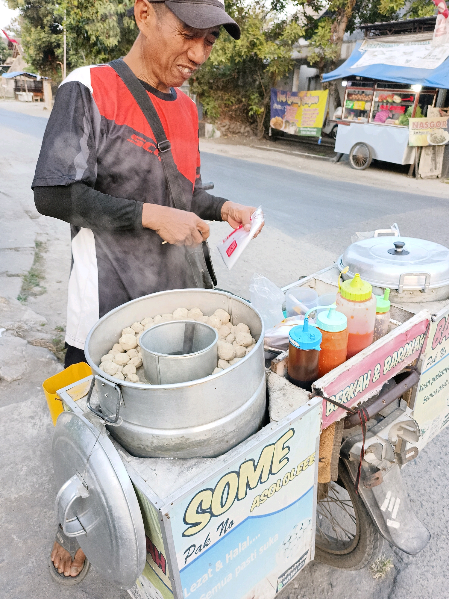 A middle-aged Indonesian man is standing on a paved street in Borangan, Indonesia, next to his food cart. The man, wearing a black and red t-shirt and a black baseball cap, is smiling as he holds a small white packet in his right hand. Steam rises from a large metal pot filled with meatballs on his cart. The cart is equipped with various condiments and a large covered pot. In the background, other food stalls and trees line the street. The scene captures a typical street food vendor in an Indonesian city.