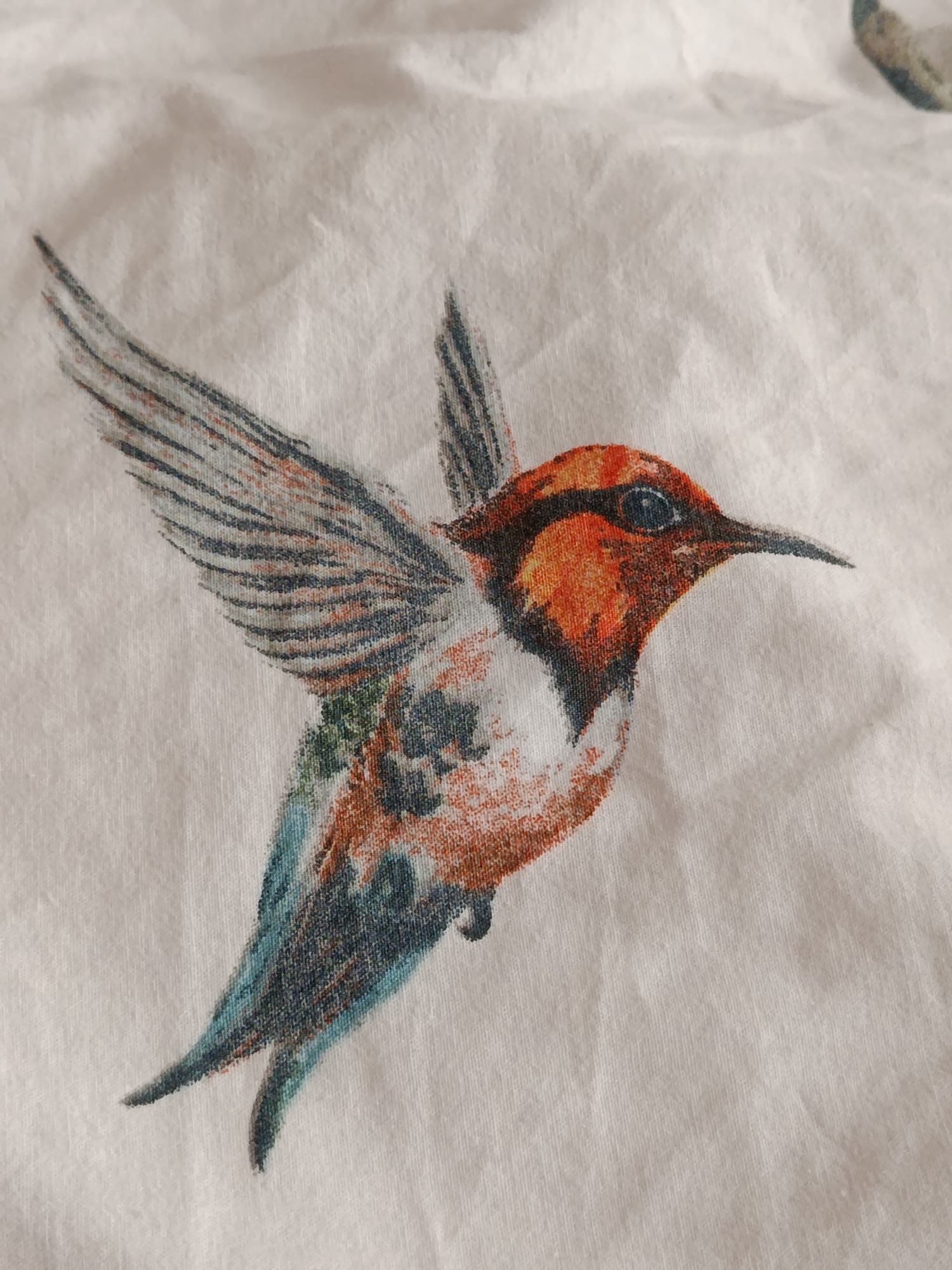 A close-up, high-angle shot captures a printed image of a hummingbird on a textured white fabric. The hummingbird is depicted in mid-flight, with its wings spread. Its body is predominantly orange and white, with iridescent green and blue on its wings and tail. The bird has a dark eye and a long, slender beak. The fabric exhibits wrinkles and a subtle weave pattern, indicating it may be a piece of clothing or home textile. No elements suggestive of the city of Svietlahorsk, Belarus, are present.