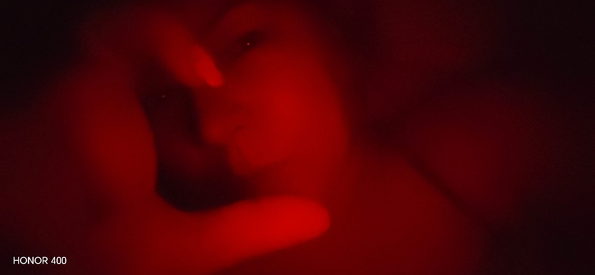 The image presents a highly abstract and dark composition, dominated by deep red hues. It appears to be a close-up, heavily blurred shot, possibly of a person. In the central area, indistinct shapes suggest a human face, with two small, bright pinpricks of light that could be reflections from eyes. A form resembling a hand or fingers is visible on the left side, seemingly touching or partially obscuring the face.
The scene is bathed in a strong, reddish light, creating deep shadows and minimizing discernible detail. The significant blur throughout the image renders specific features and expressions unidentifiable, contributing to an enigmatic and moody atmosphere. The overall impression is one of intimacy and mystery, conveyed through the low-light conditions and the intense monochromatic red coloring. There are no clear environmental or location cues within the image itself. In the bottom left corner, the text "HONOR 400" is visible, likely a watermark.
