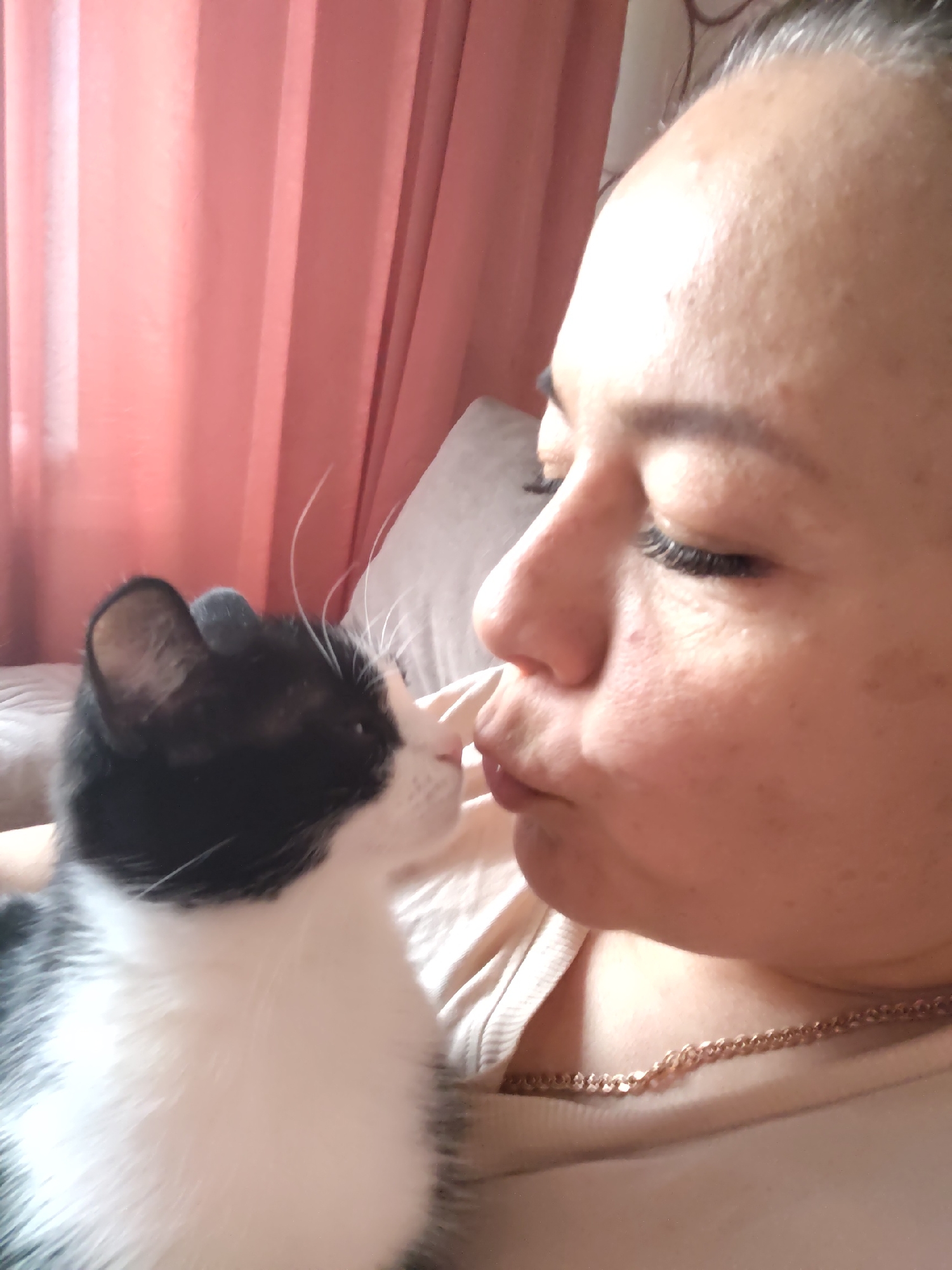 A woman with closed eyes is seen in close proximity to a black and white cat. The woman's face is turned towards the cat, and their noses appear to be touching. She is wearing a light-colored garment and a gold chain necklace. The cat has white whiskers and primarily white fur on its body, with black fur covering its head and ears. Behind the woman are pink curtains. The location is unknown.