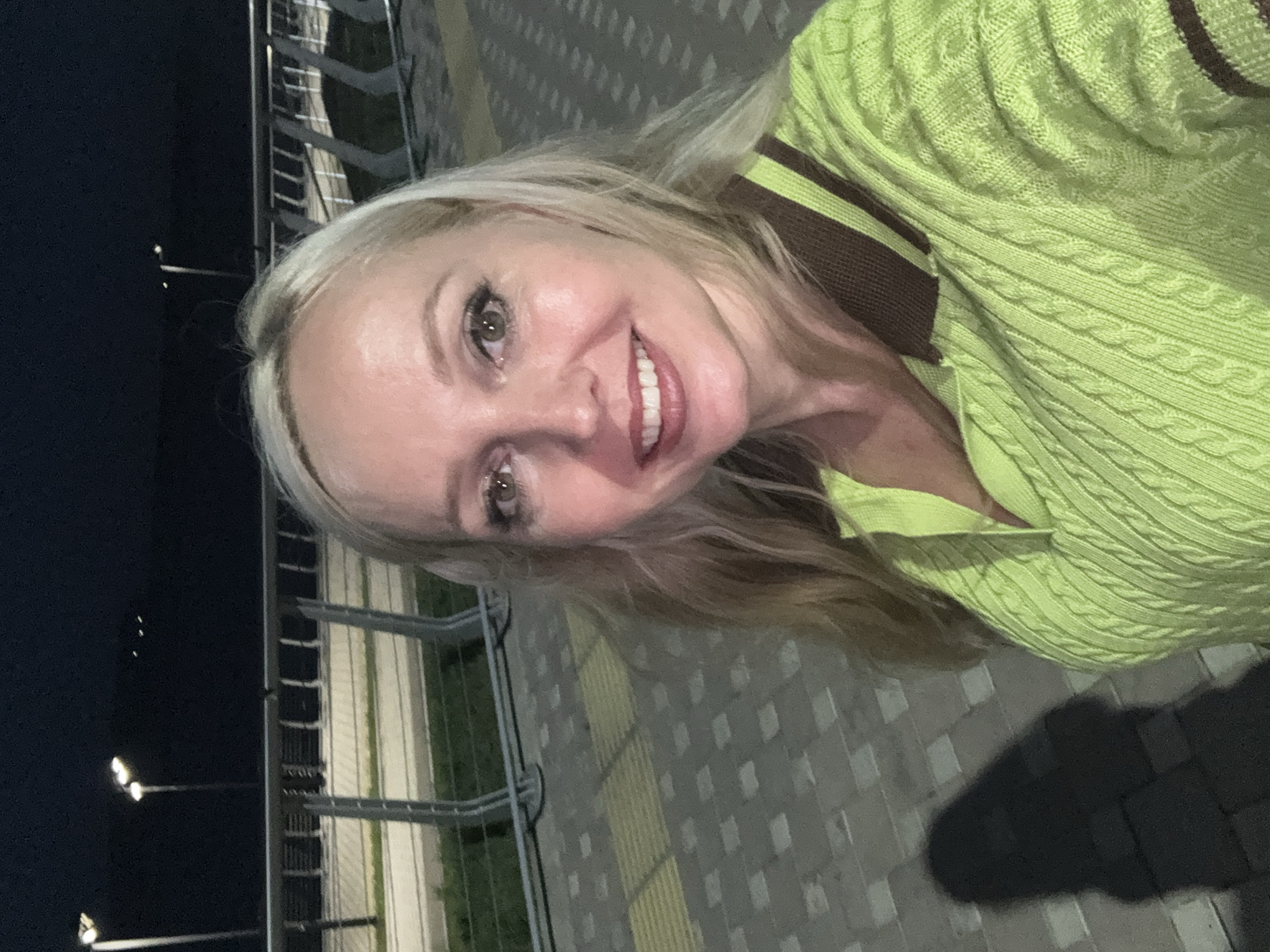 In Komsomolsk-on-Amur, Russia, at night, a fair-skinned woman with blonde hair takes a selfie outdoors. She wears a bright lime green cable-knit sweater with a brown collar and trim. Her eyes are open, and she is smiling, exposing her teeth. Behind her, a metal railing with vertical supports is visible, bordering a paved walkway and an area with green grass. Streetlights illuminate the scene, casting a shadow of the woman on the patterned pavement.