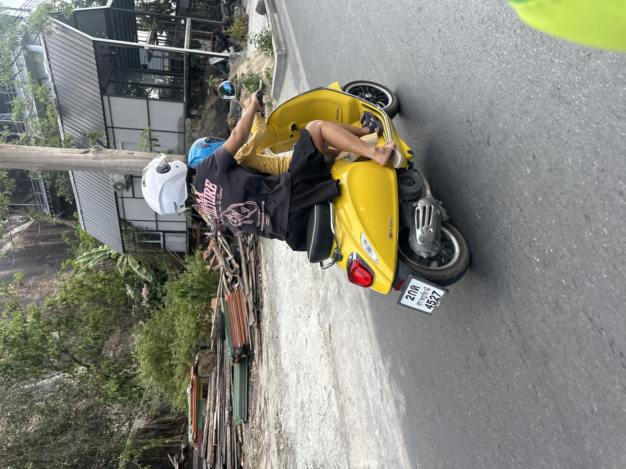 Two individuals are captured riding a vibrant yellow Vespa Sprint scooter on a paved road, likely in Baan Chaweng Noi, Thailand, given the local context and visible Thai script. The image is oriented sideways, with the scooter appearing to ride vertically.
The driver, wearing a white LS2 helmet and a black t-shirt with black shorts, is a male holding the handlebars. His black t-shirt features white text including "NURE," "That The Queen," "B+2567," and "BECAUSE UNIFORM AMATEURS." Behind him, a passenger, partially visible and wearing a yellow top and pants, rides without a helmet. This passenger has their bare feet, clad in light-colored flip-flops, casually resting on the scooter's front floorboard. A blue helmet can be seen strapped to the back of the scooter, perhaps belonging to the passenger or a spare.
The setting is an outdoor, daytime scene with clear weather. The road appears to be asphalt, flanked by a dirt or gravel path on one side. To the left (in the corrected orientation), there's a mixture of lush green foliage, large trees, and a prominent grey rock formation or cliff. On the right, various buildings with metal roofs and glass windows are visible, alongside piles of what appears to be construction debris or stacked wooden planks and colorful corrugated sheets. The overall atmosphere suggests a casual ride in a somewhat built-up yet green and rocky area.
Notable text details include "Sprint" and "ABS" on the scooter body. The license plate clearly displays Thai script "2กด," "สุราษฎร์ธานี" (Surat Thani), and the numbers "4527."