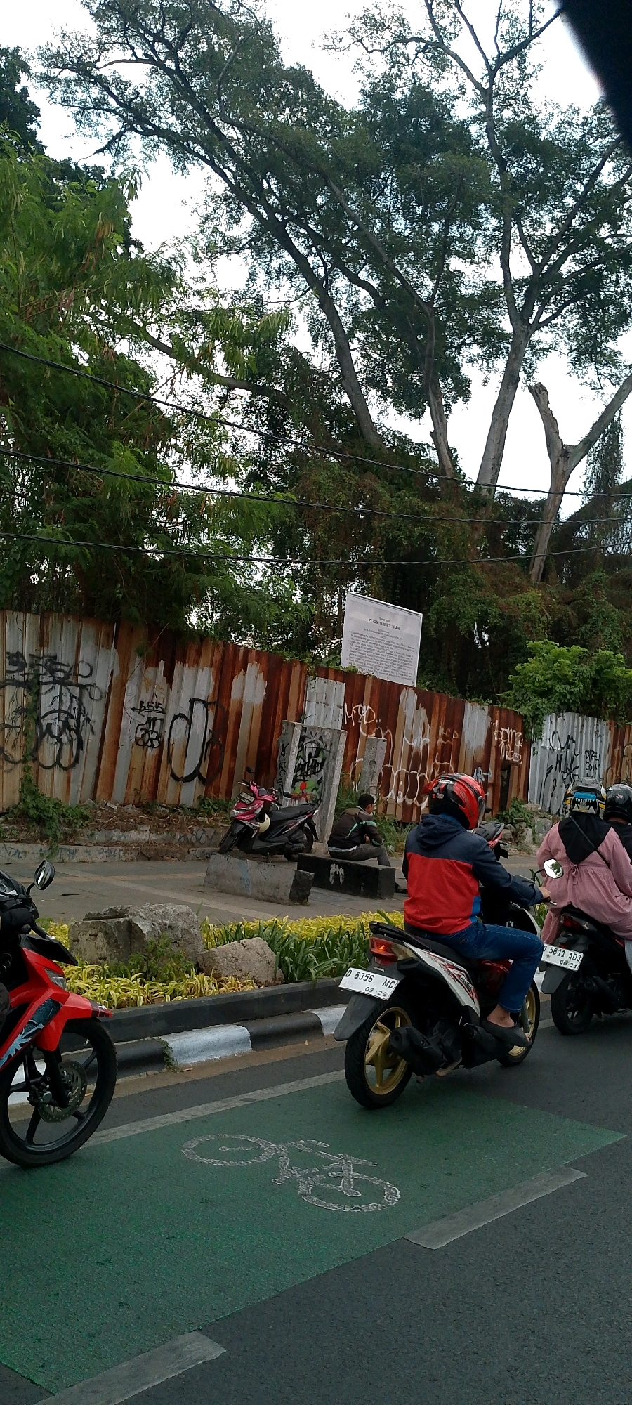 The image depicts a street scene in Bandung City, Indonesia. In the foreground, a green lane with a bicycle symbol painted on it is visible, indicating a bike lane. A red motorcycle is on the left, and two other motorcycles are further down the road, one with a rider in a red and blue jacket and the other with a rider in a pink outfit. In the background, a rusty metal fence with graffiti covers a section of the landscape, and large trees with lush green foliage dominate the upper part of the frame. The sky is overcast. There are a few stones and some yellow plants planted along the side of the road. A sign is attached to the fence in the background.