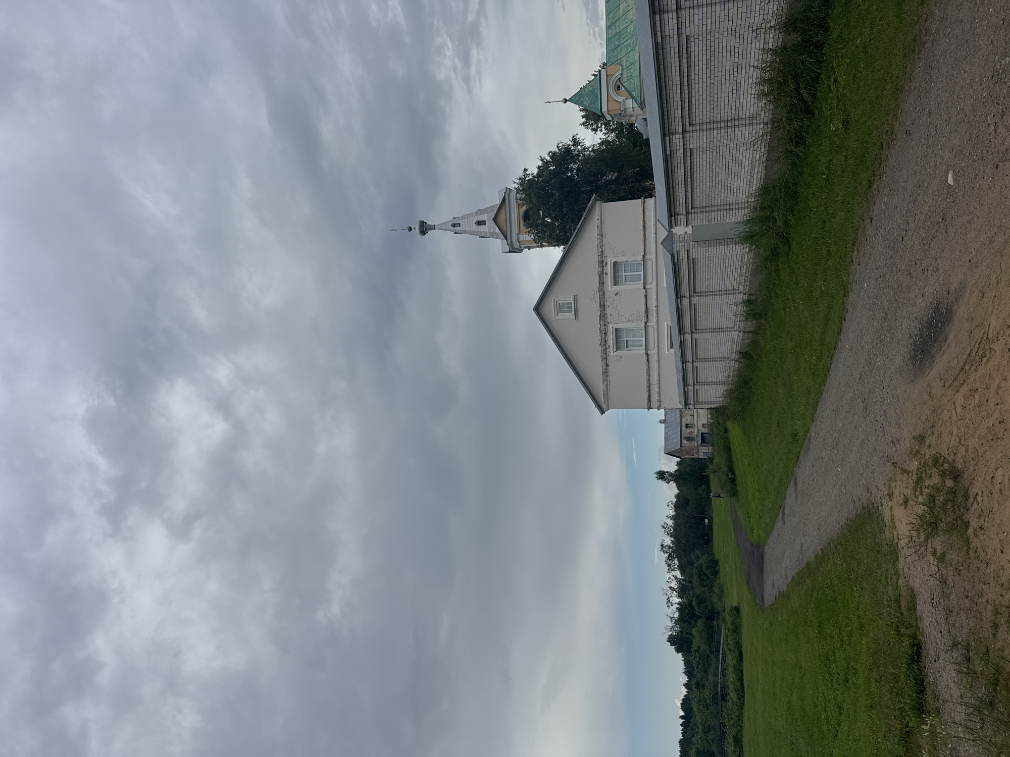 The image captures a view of a pathway leading towards a walled compound in the city of Рассвет, Russia. The sky is overcast with dense, gray clouds. To the right, a white brick wall borders the property, behind which stands a pale pink building with white-trimmed windows. Beyond this building, a prominent bell tower with a white spire and yellow accents rises into the sky, partially obscured by lush green trees. To the left of the pathway, a vast expanse of green grass stretches towards a line of dense trees and foliage. In the distance, a pedestrian can be seen walking along the pathway, and a rustic wooden bridge is visible crossing a body of water within the greenery.
